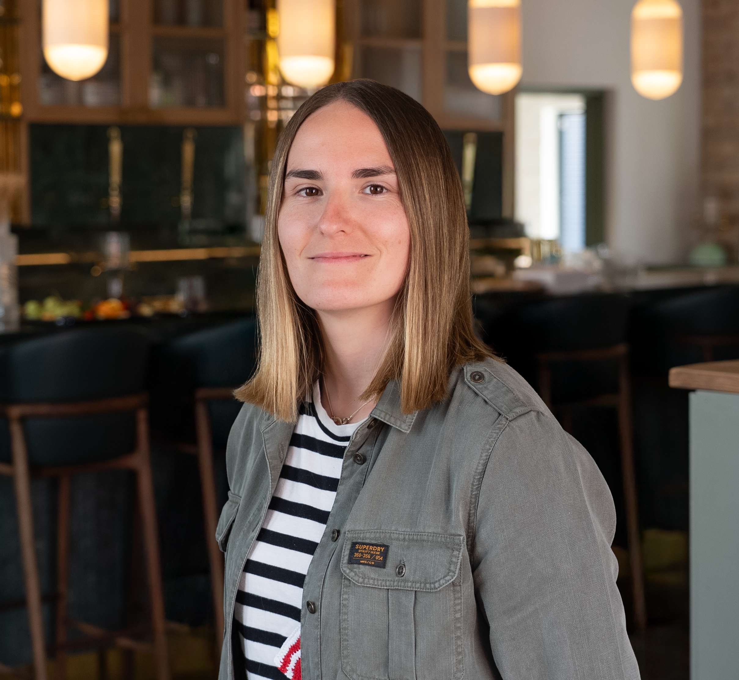 Lucy Brown, a young woman with shoulder-length light brown hair, wearing a gray jacket over a black and white striped shirt, is smiling softly in a modern café or restaurant setting with warm lighting and wooden accents.