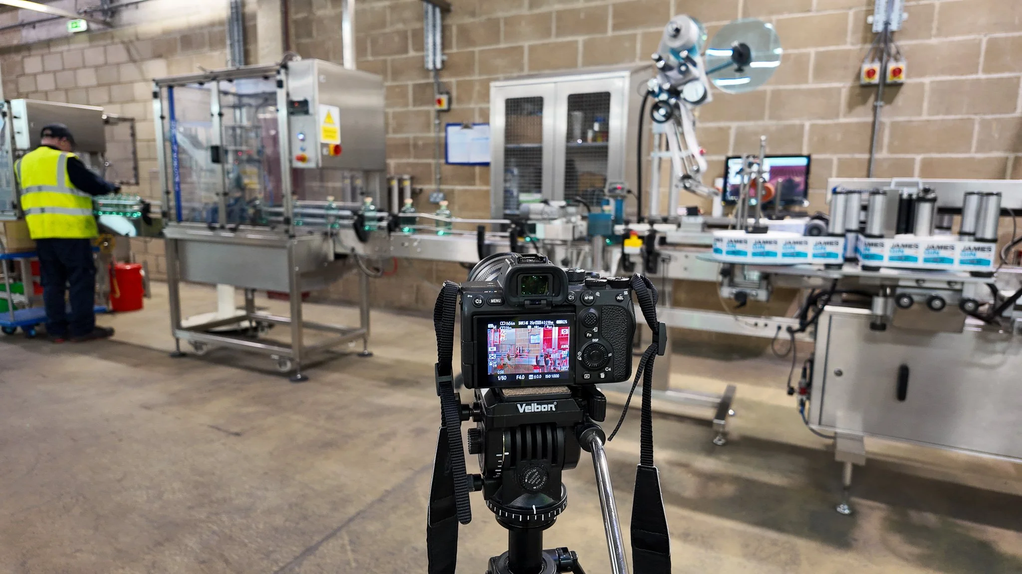 Camera capturing an industrial manufacturing setting with a worker in a high visibility vest, machinery, and conveyor belts for packaging or production.