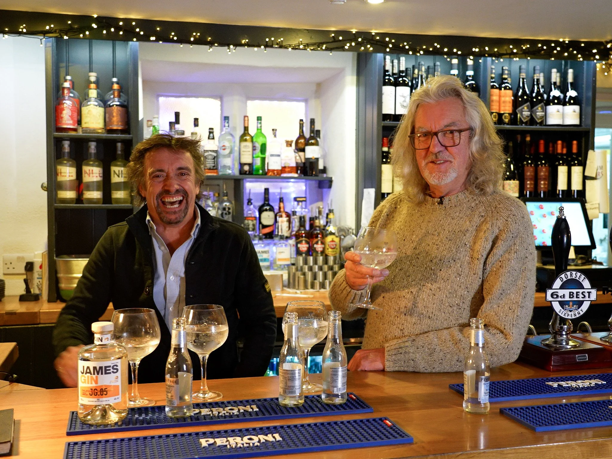 James May and Richard Hammond standing at a bar, smiling and holding drinks, with shelves of liquor bottles in the background and string lights overhead.
