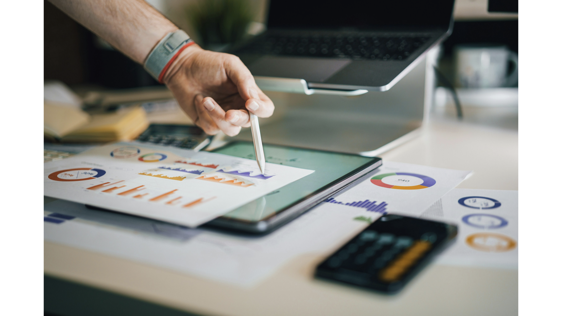A person using a stylus on a tablet with financial charts and graphs, with a laptop, calculator, papers with colorful data visualizations, and a smartphone on a desk. Analysing data to increase drink company marketing and sales performance.