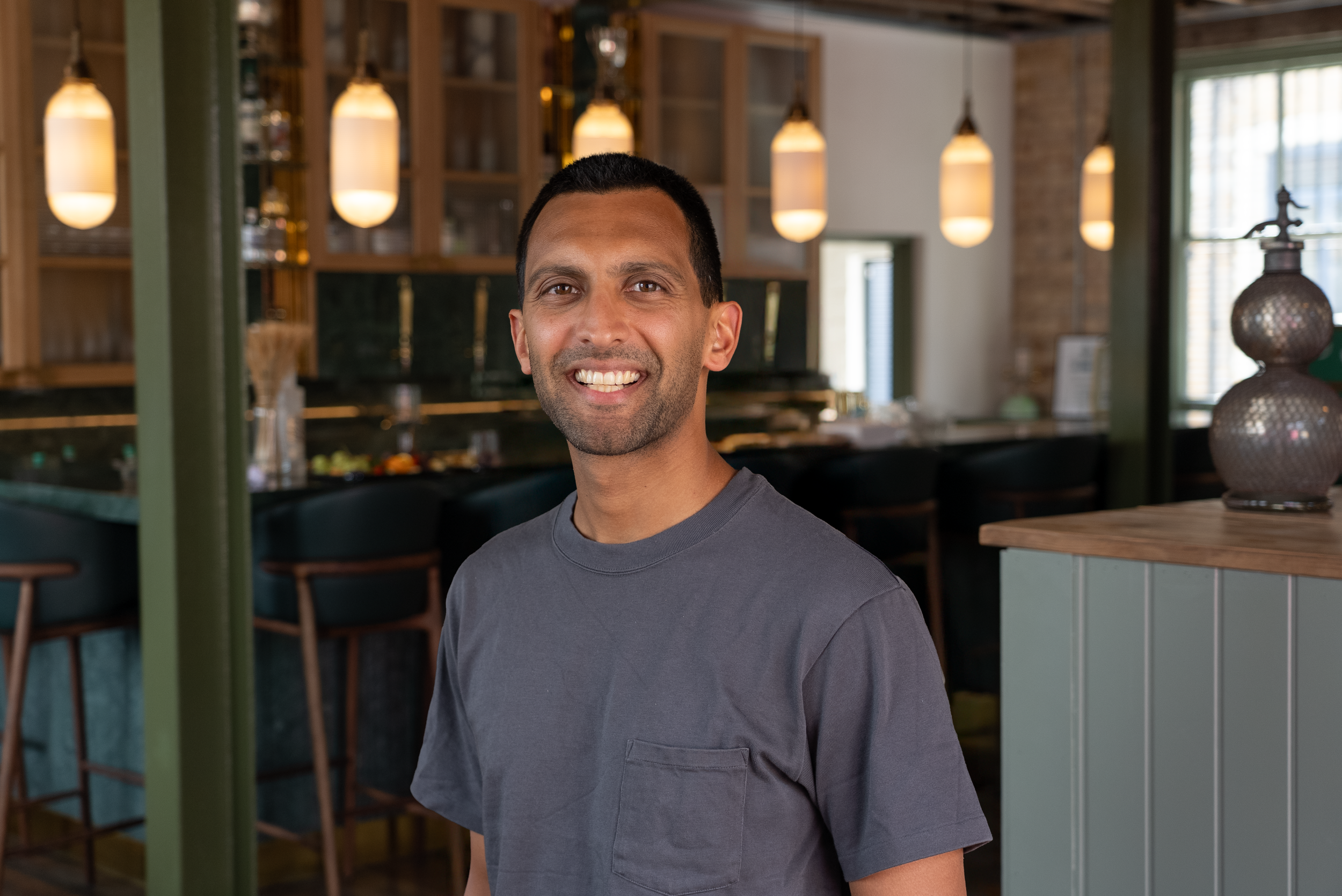 Vishal Patel, co-founder of Straight Shot, standing inside a restaurant with hanging pendant lights, a bar counter, and decorative vases.