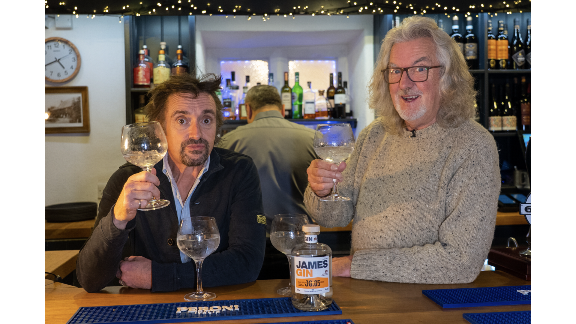 James May and Richard Hammond at a bar, raising glasses of gin, with a bottle of James Gin on the bar in front of them. The bar is decorated with shelves of liquor bottles, and there are lights above the shelves.
