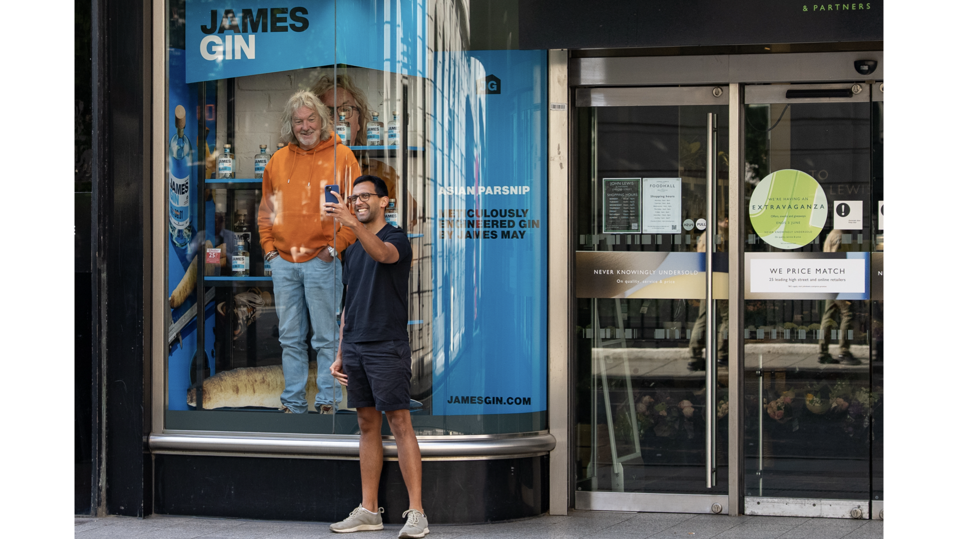 Two men taking a selfie outside a storefront with promotional posters. One man is James May founder of James Gin. The other man is taking a selfie with him. James May is in the John Lewis store window promoting James Gin.