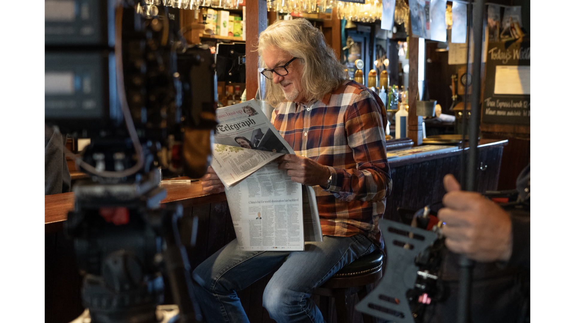 James May sitting on a barstool reading a newspaper in a dimly lit pub or bar during the filming of social media and youtube content to promote his drinks brand, James Gin.