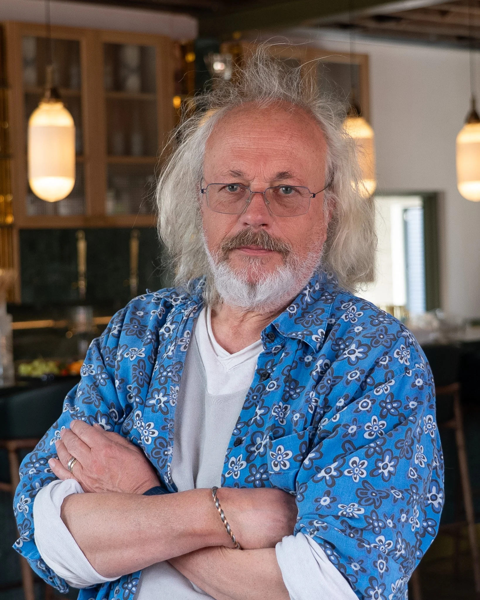 A man with long, gray, messy hair and a beard stands with arms crossed in a cozy, well-lit interior. He's wearing glasses, a white shirt, and a blue floral-patterned shirt. The background features warm lighting fixtures and a kitchen or dining area.