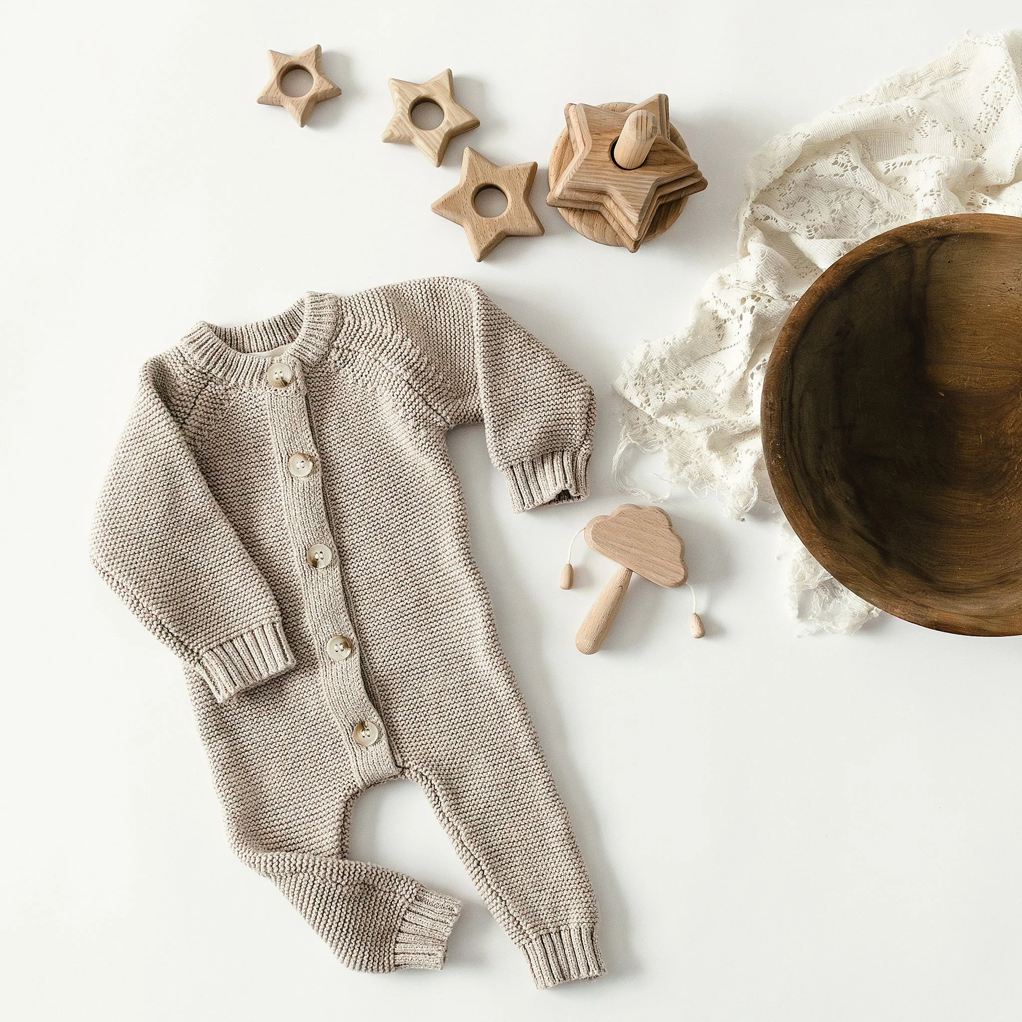 A beige knitted baby onesie with buttons, wooden star-shaped toys, a wooden mushroom-shaped teething toy, lace fabric, a wooden bowl, and a black and white patterned cloth on a white surface for a flat lay photography session in Naperville, IL.