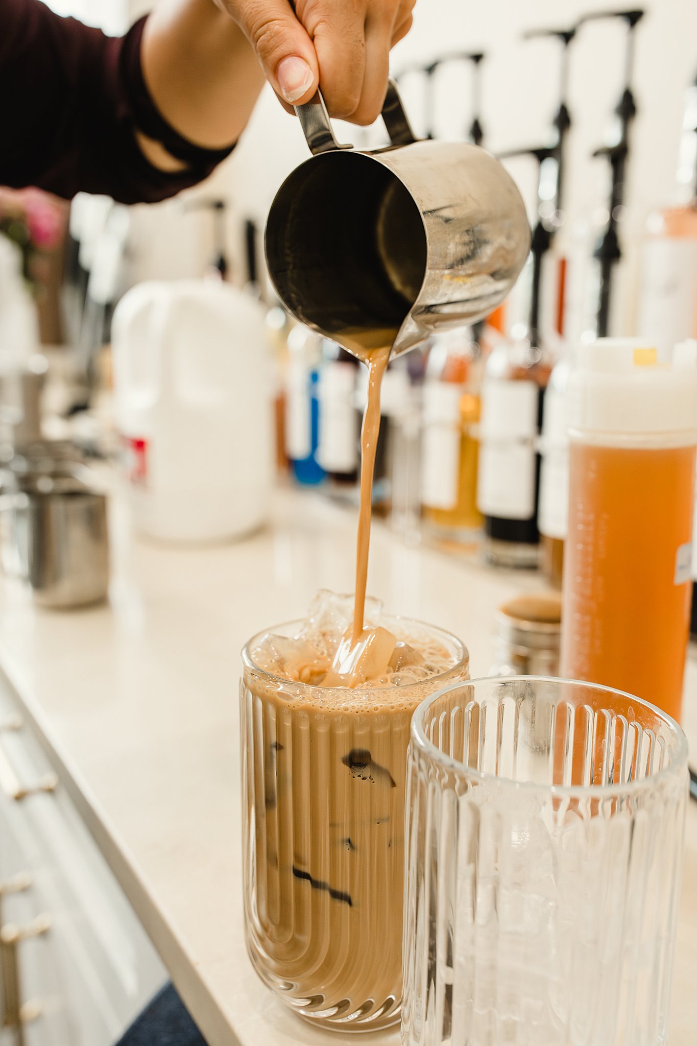 Pouring iced coffee into a glass from a metal pitcher with blurred kitchen items in the background for business branding photography.