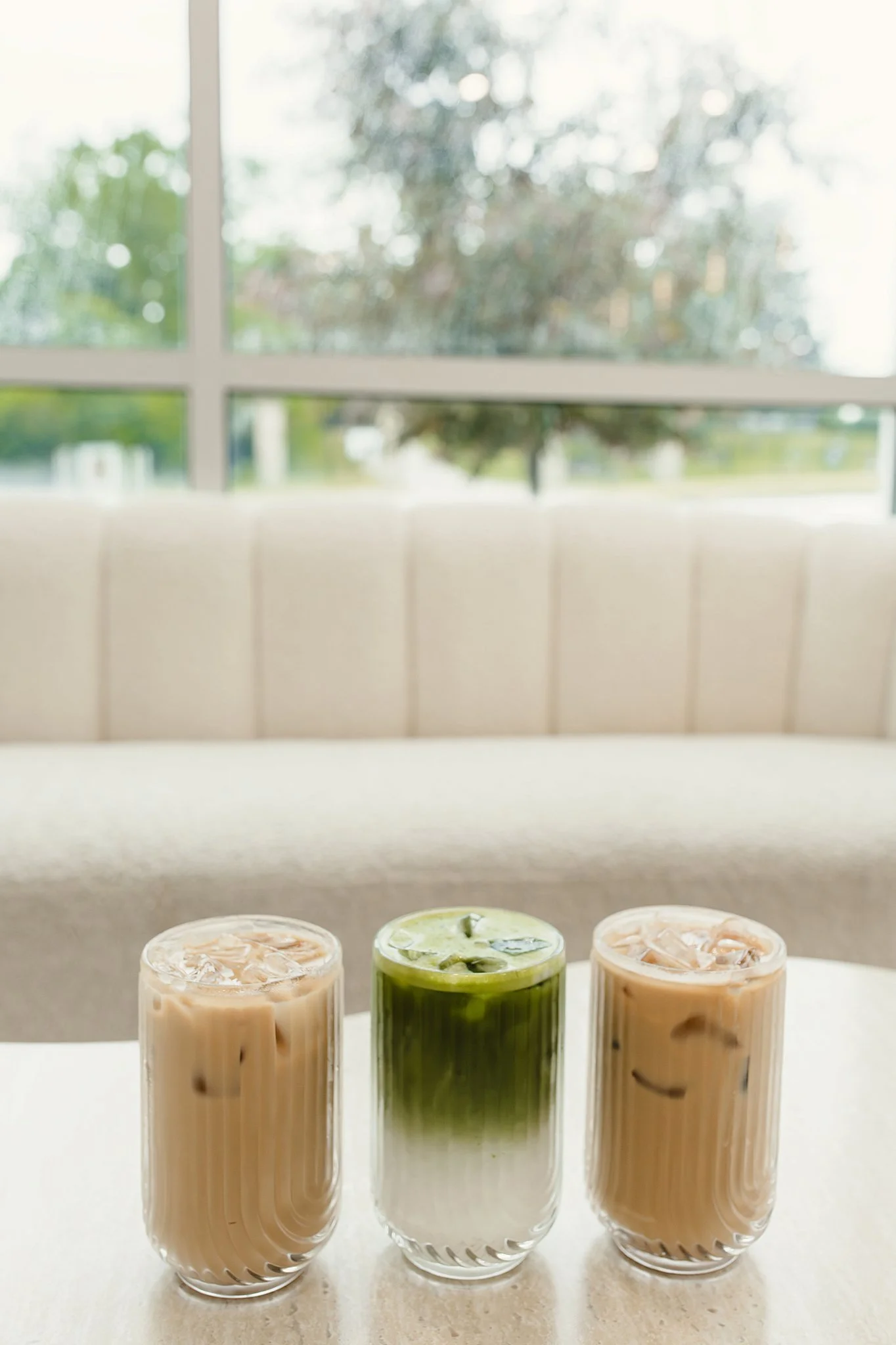 Three glasses of iced coffee or tea with different flavors, placed on a white surface with a window showing trees in the background for coffee shop photography.