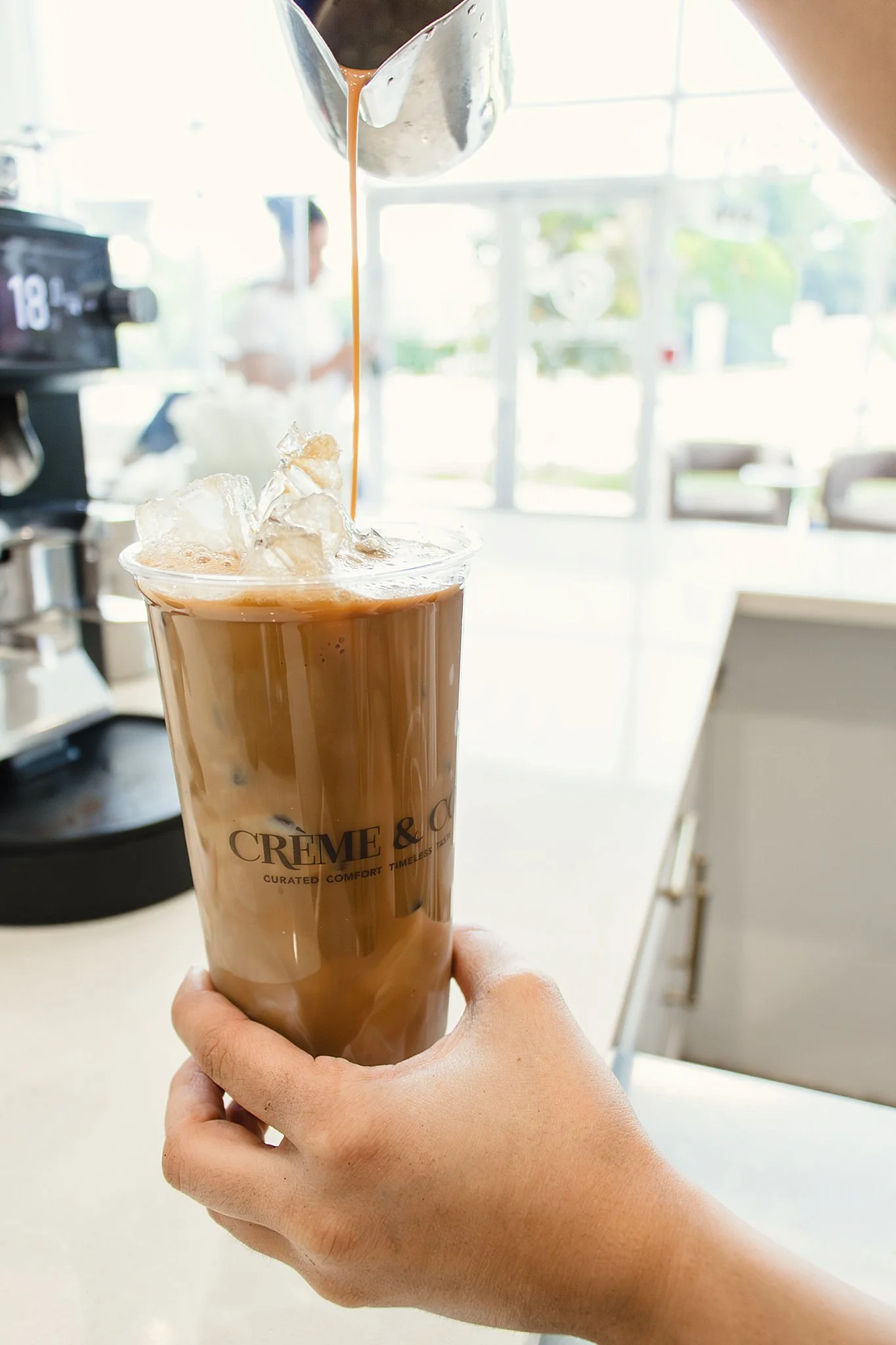 A hand holding a tall glass of iced coffee with a straw, on a light-colored table in a bright cafe. The glass has the words 'CREME & CO' printed on it with branding photographers near Naperville, IL.