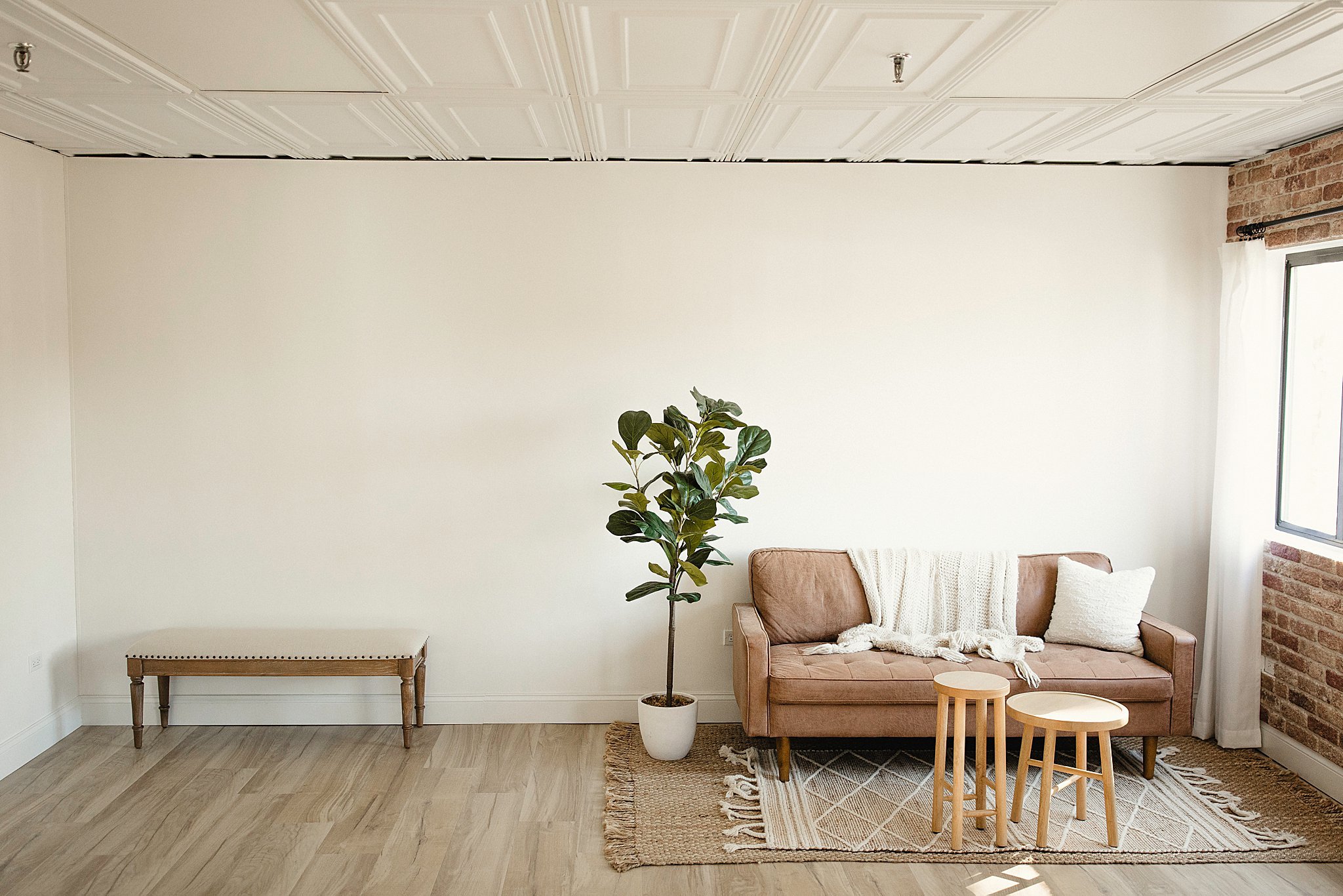 Minimalist living room with a tan sofa, a white throw blanket and pillows, a potted plant, two small wooden stools, a woven rug, and a large window with white curtains at the Ally and B Photography studio in Naperville, IL.