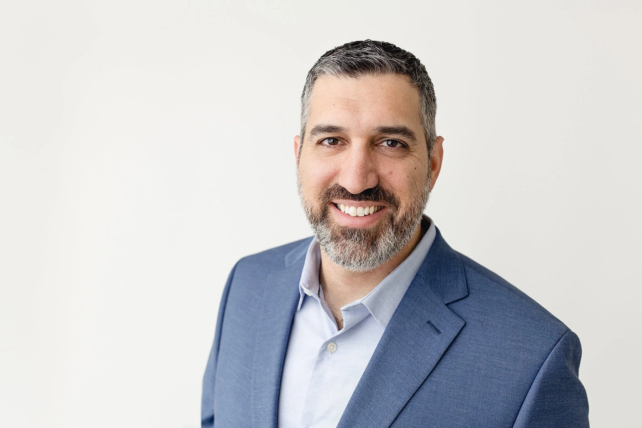Portrait of a middle-aged man with gray hair and beard, wearing a blue suit and light blue shirt, smiling against a plain white background during headshot photography with Ally and B Photography.