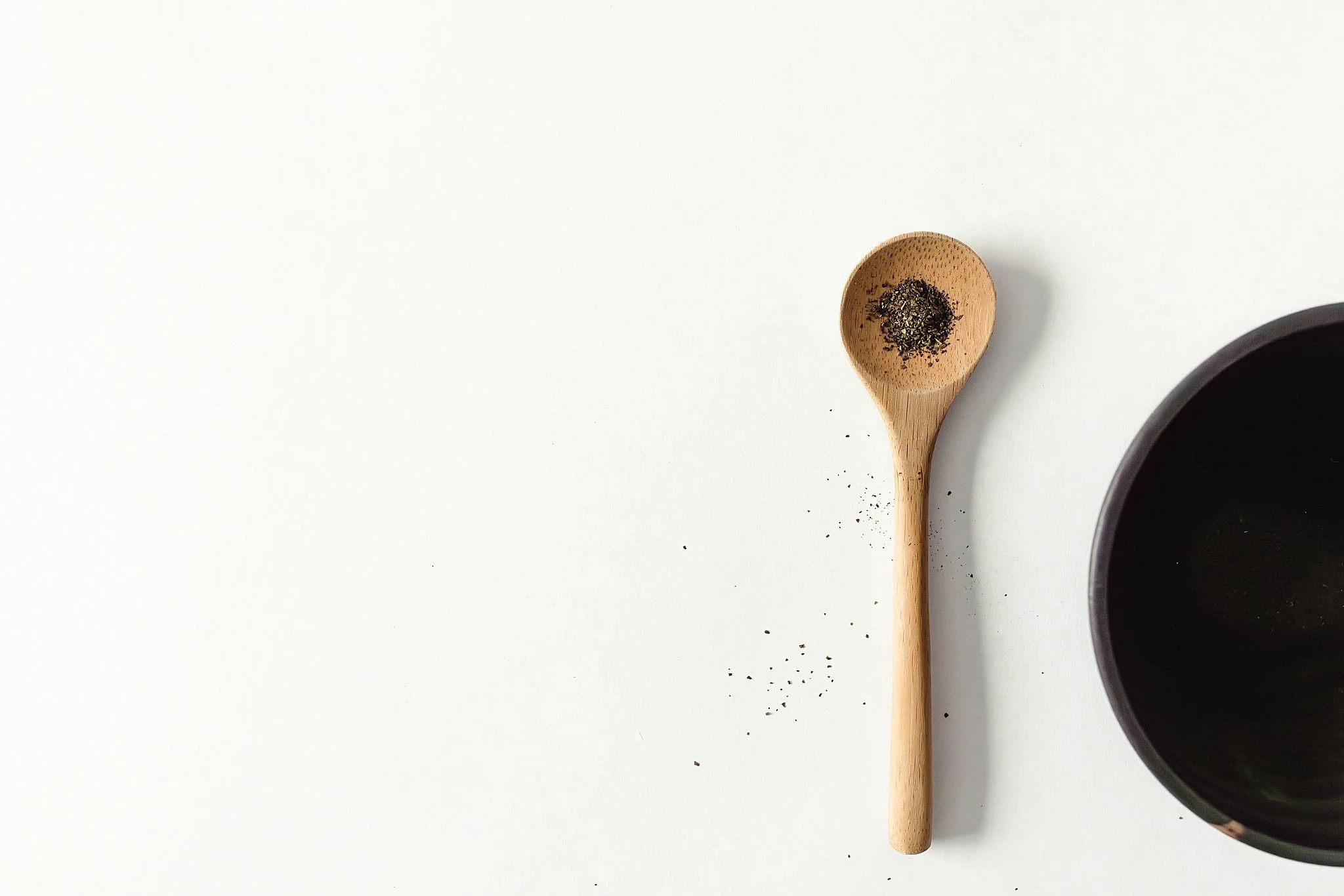 A wooden spoon with black pepper inside, some pepper scattered below, and part of a black bowl visible on the right side, all on a white surface for product photos near me.