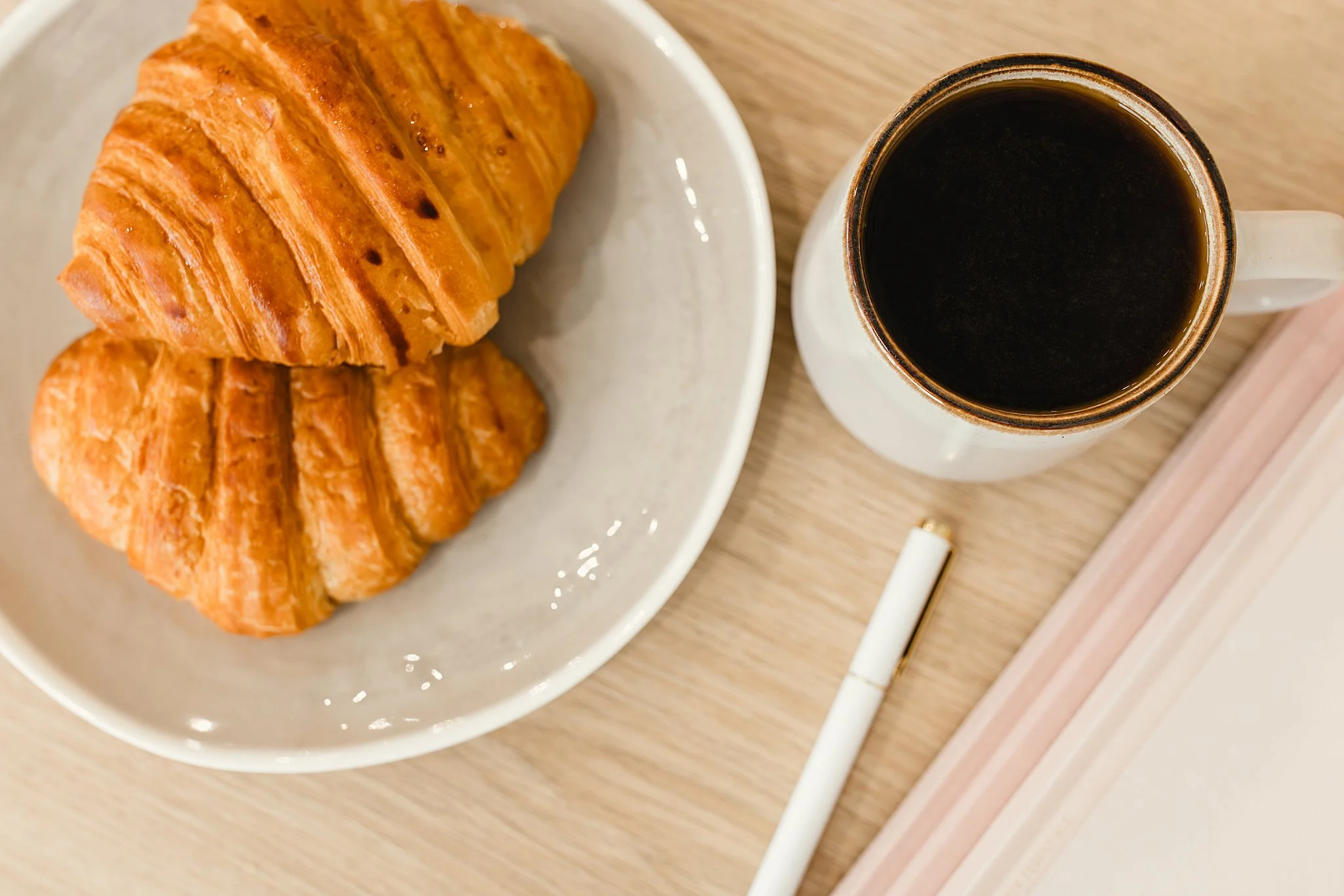A white plate with two golden brown croissants, and a white mug filled with black coffee on a wooden table with a pink tray for food and drink branding photography Naperville.