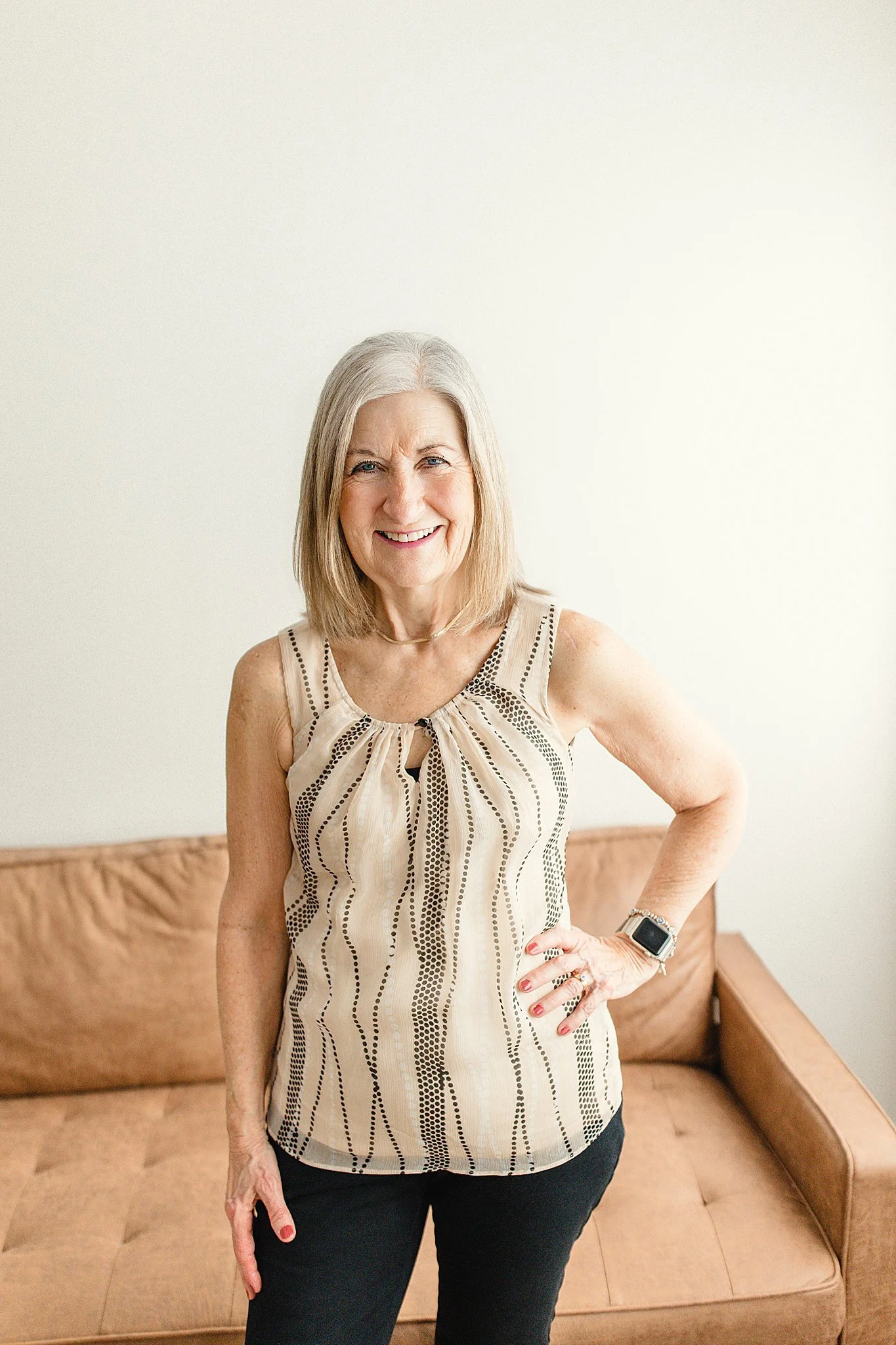 An elderly woman with gray hair smiling, standing in front of a beige couch and plain white wall, wearing a sleeveless patterned beige top and black pants with brand photographers near me.
