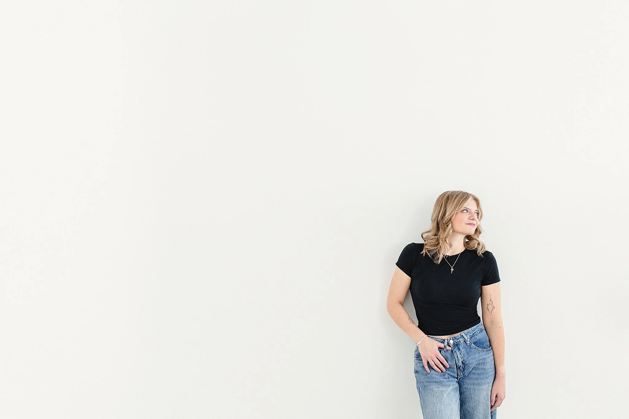 A woman with blonde hair wearing a black t-shirt and blue jeans standing against a plain white wall, looking to her right for a personal branding session in Naperville, IL.