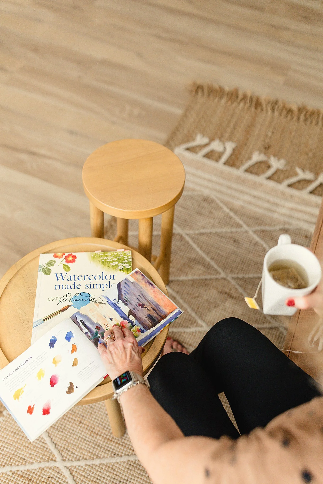 Person sitting on a chair reading a watercolor book with a color palette, holding a tea mug, with a small round wooden table nearby, on a rug and wooden floor during branding photography near me.