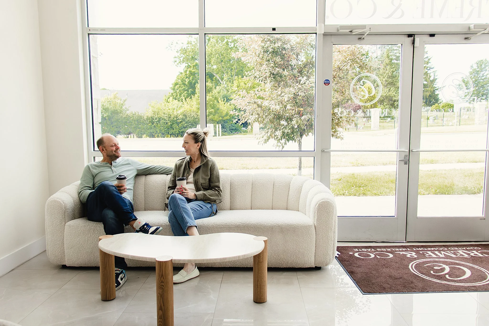 Two people sitting on a white couch near large window with trees outside, holding coffee cups, smiling at each other with brand photoshoot near me.