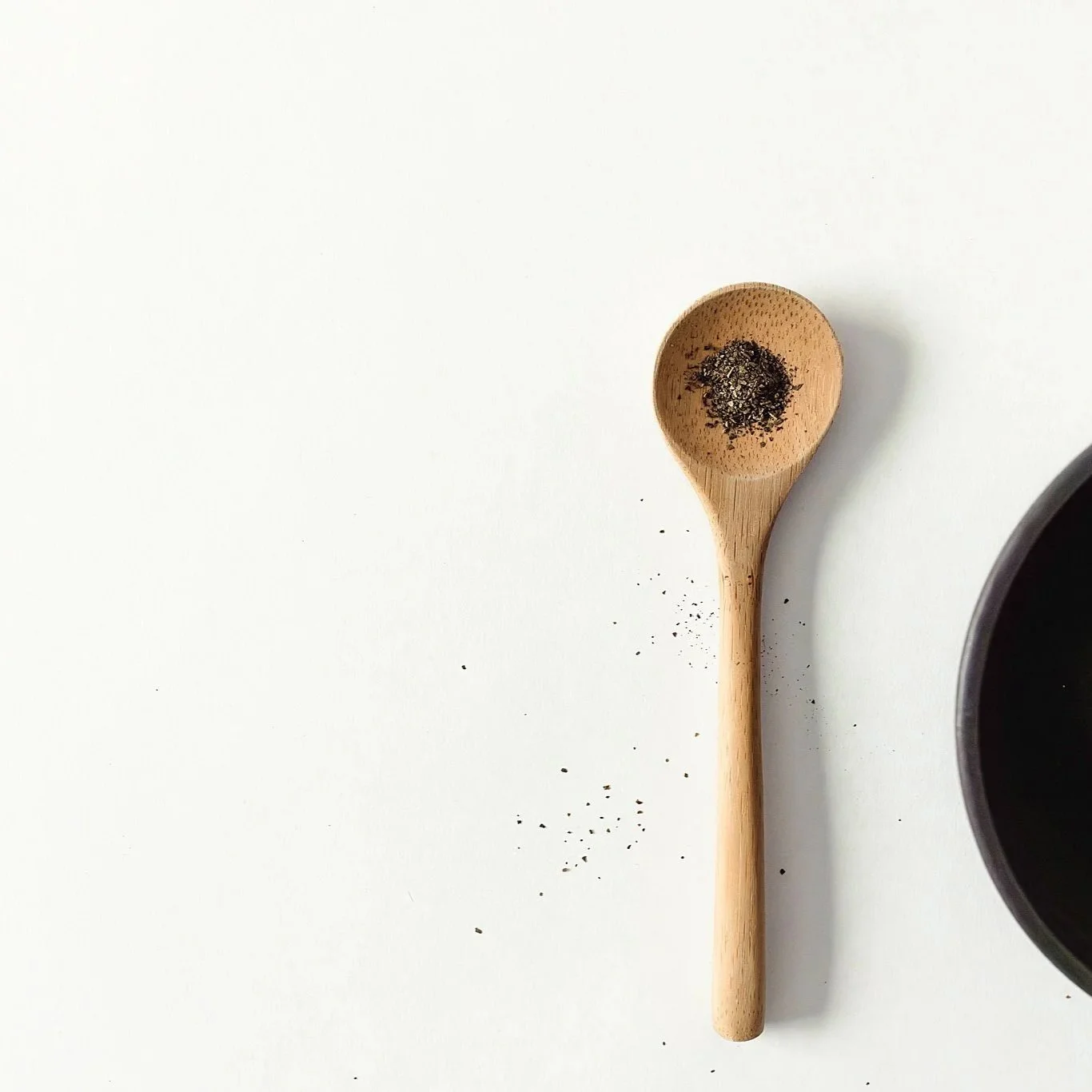 A wooden spoon with black pepper powder on top, placed next to a black container on a white surface with some pepper scattered around for a product photography photo session.
