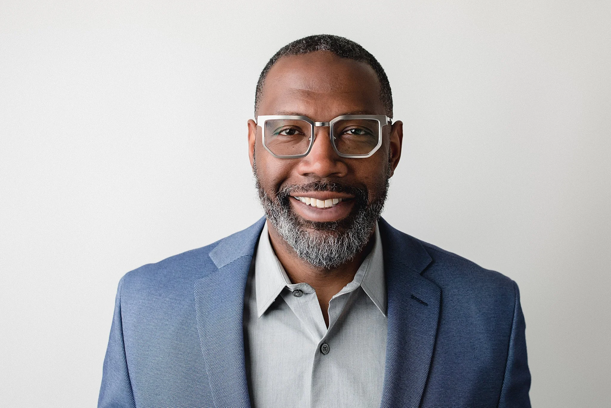 A smiling man wearing glasses and a blue blazer with a light grey shirt, standing against a plain white background during a Naperville headshot photography session.