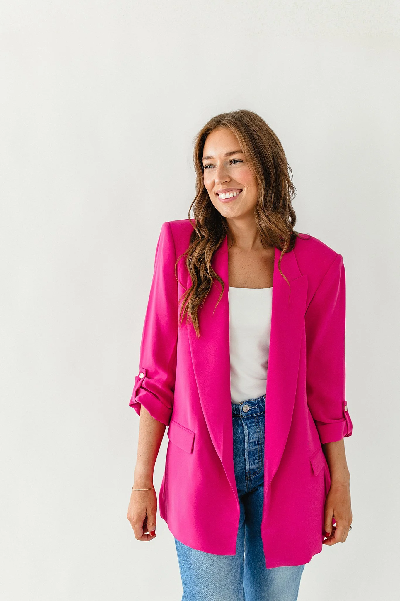 A woman with long wavy brown hair wearing a vibrant pink blazer, white top, and blue jeans, smiling against a plain white wall during a branding session near me.