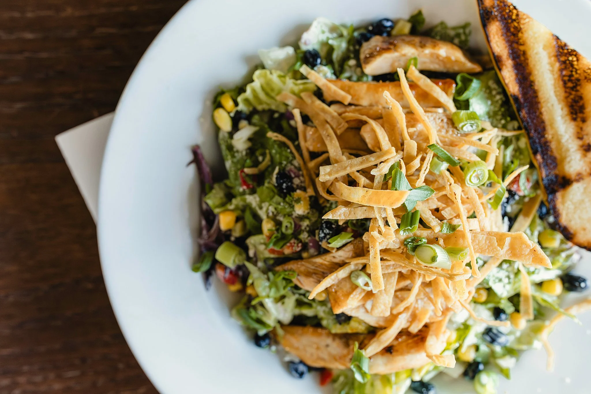A salad on a white plate with shredded chicken, green onions, corn, blackberries, and a slice of toasted bread during a bar restaurant commercial photography session for food photography of menu items.