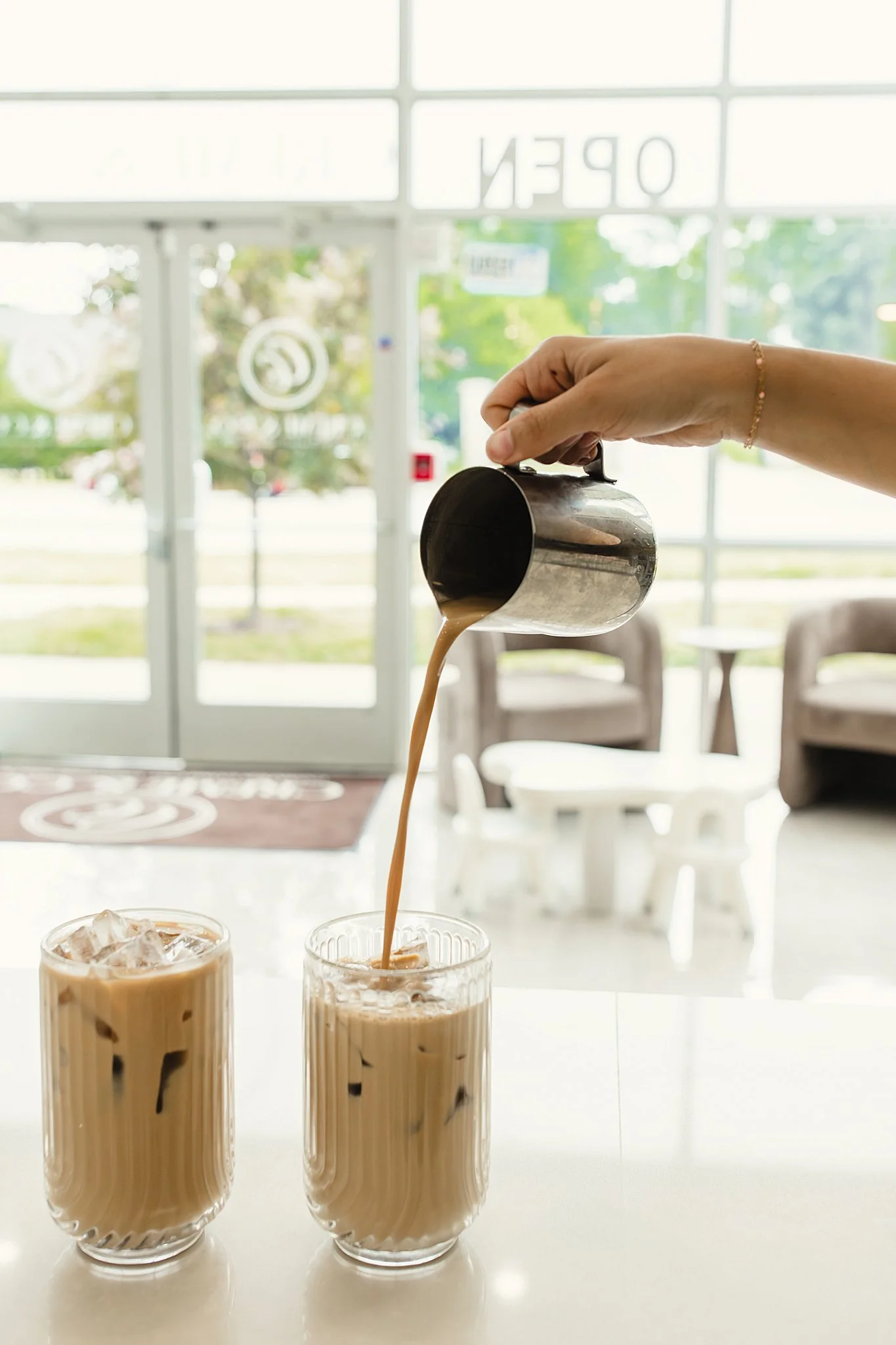 Three glasses of iced coffee with milk on a white surface, with a hand pouring coffee from a metal pitcher into one of the glasses, inside a bright cafe with glass doors and green trees outside for brand photography Naperville.