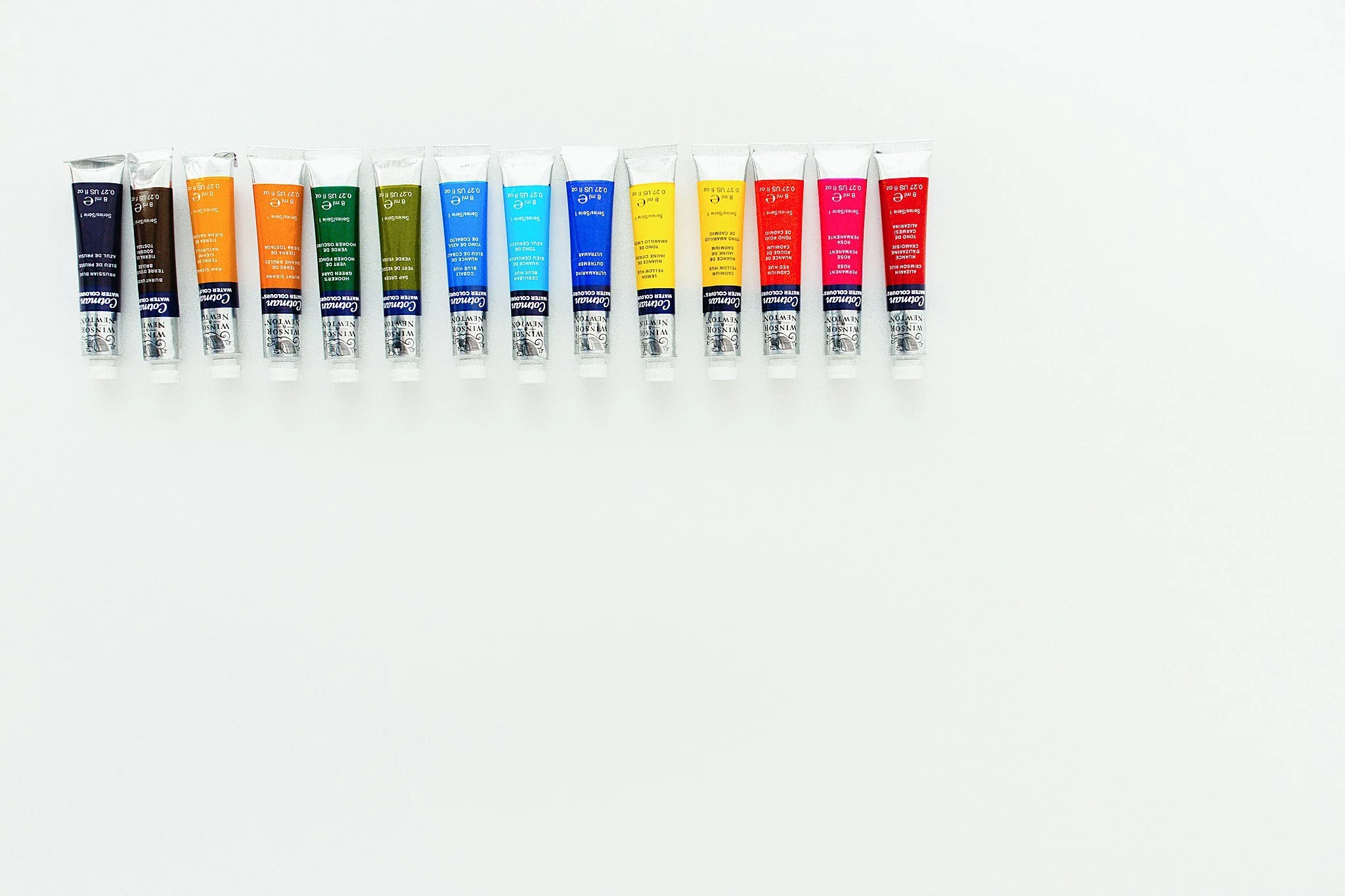 Set of 12 watercolor tubes arranged in a row on a white background, each labeled with different colors for flay lay photographers near me.