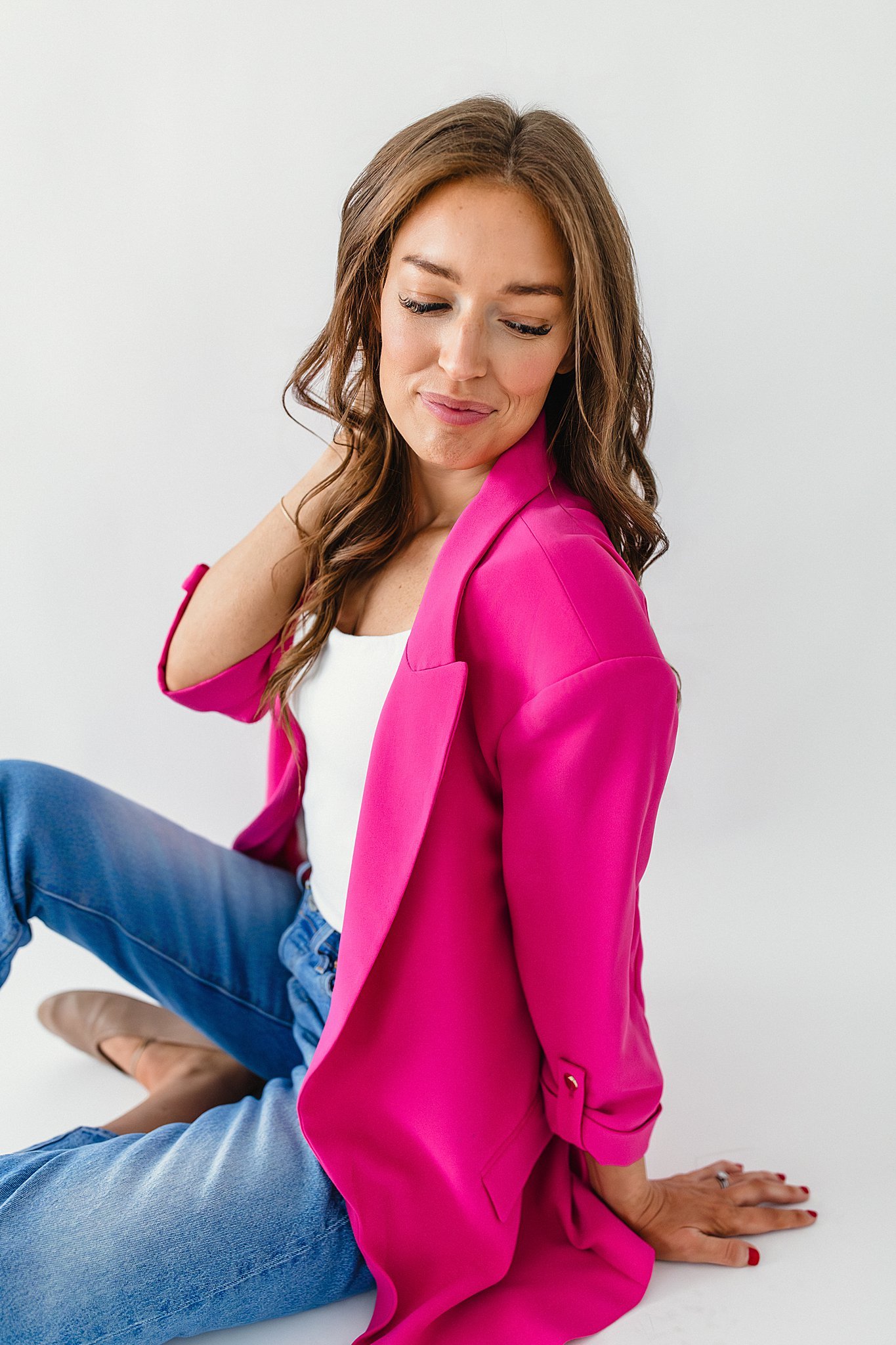 A woman with brown hair wearing a bright pink blazer, white top, and blue jeans, sitting on the floor with a white background for branding photos near Naperville, IL.