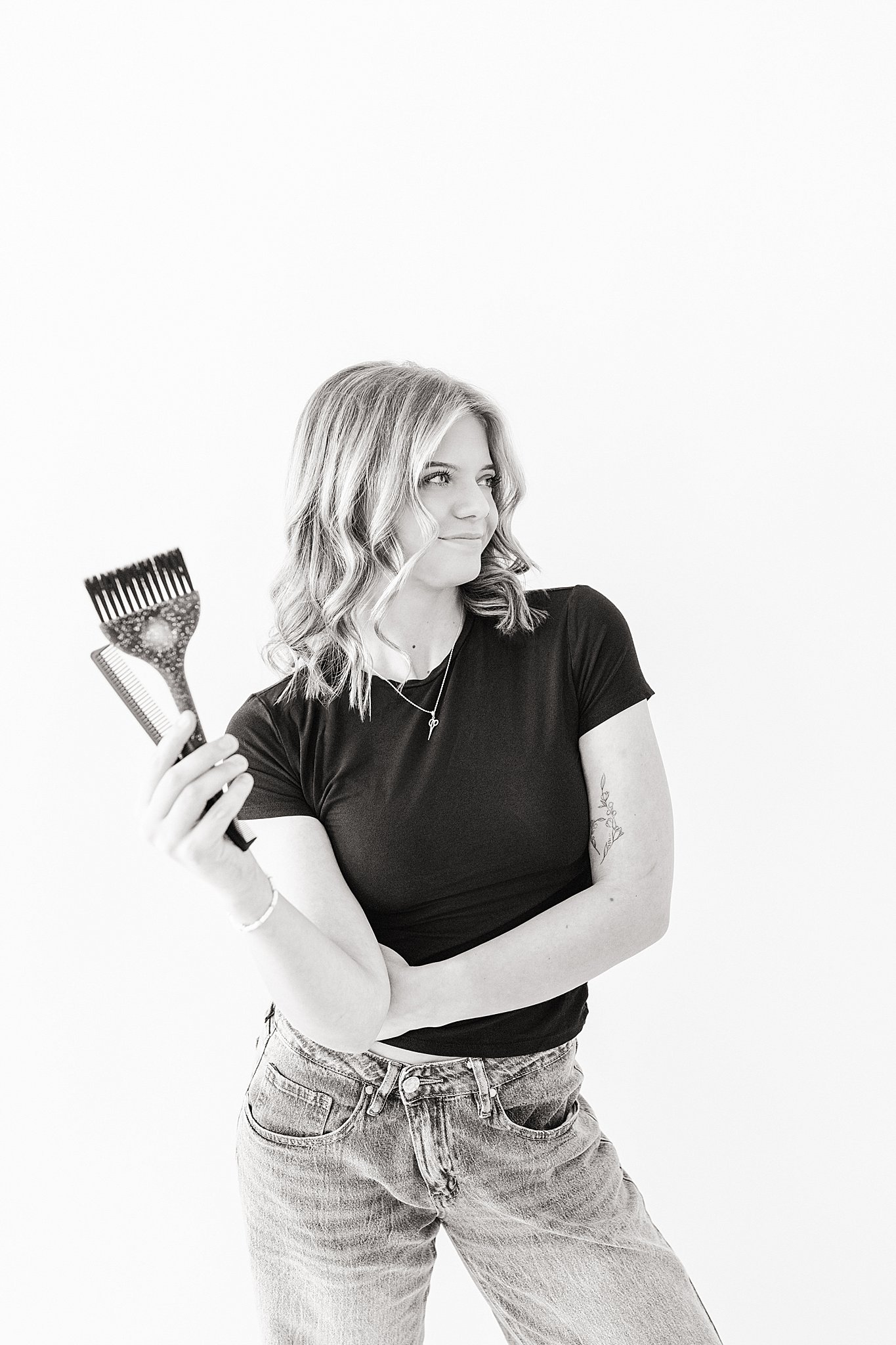 A woman with wavy shoulder-length hair and tattoos on her arm, dressed in a black t-shirt and jeans, holding a wide-tooth comb in her right hand, standing against a plain white background during branding photos in Naperville, IL.