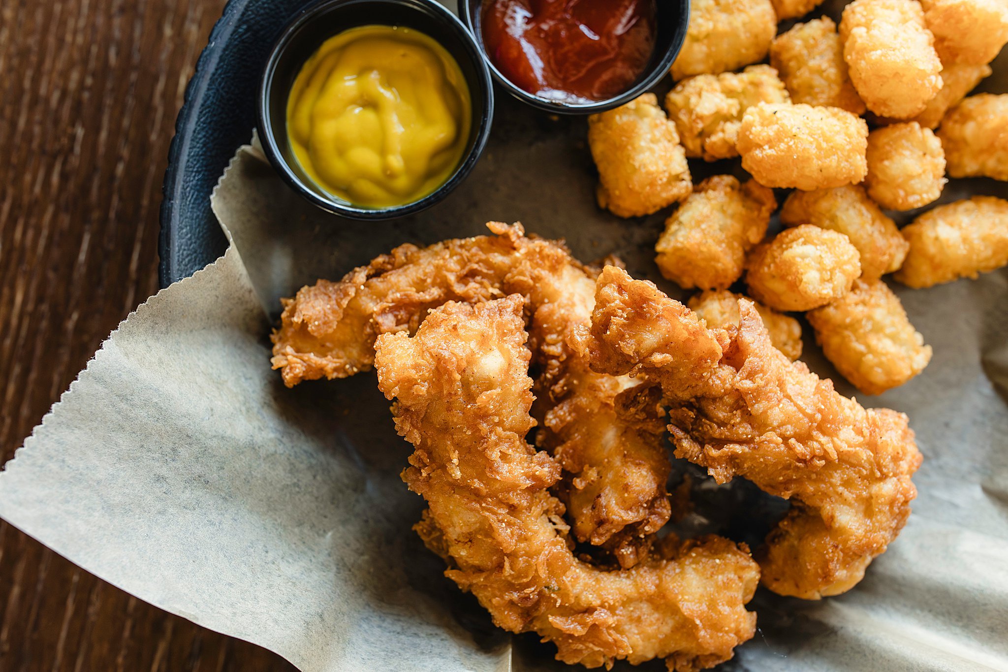 Basket with fried chicken, tater tots, and condiments including mustard and ketchup with food photographers near me.