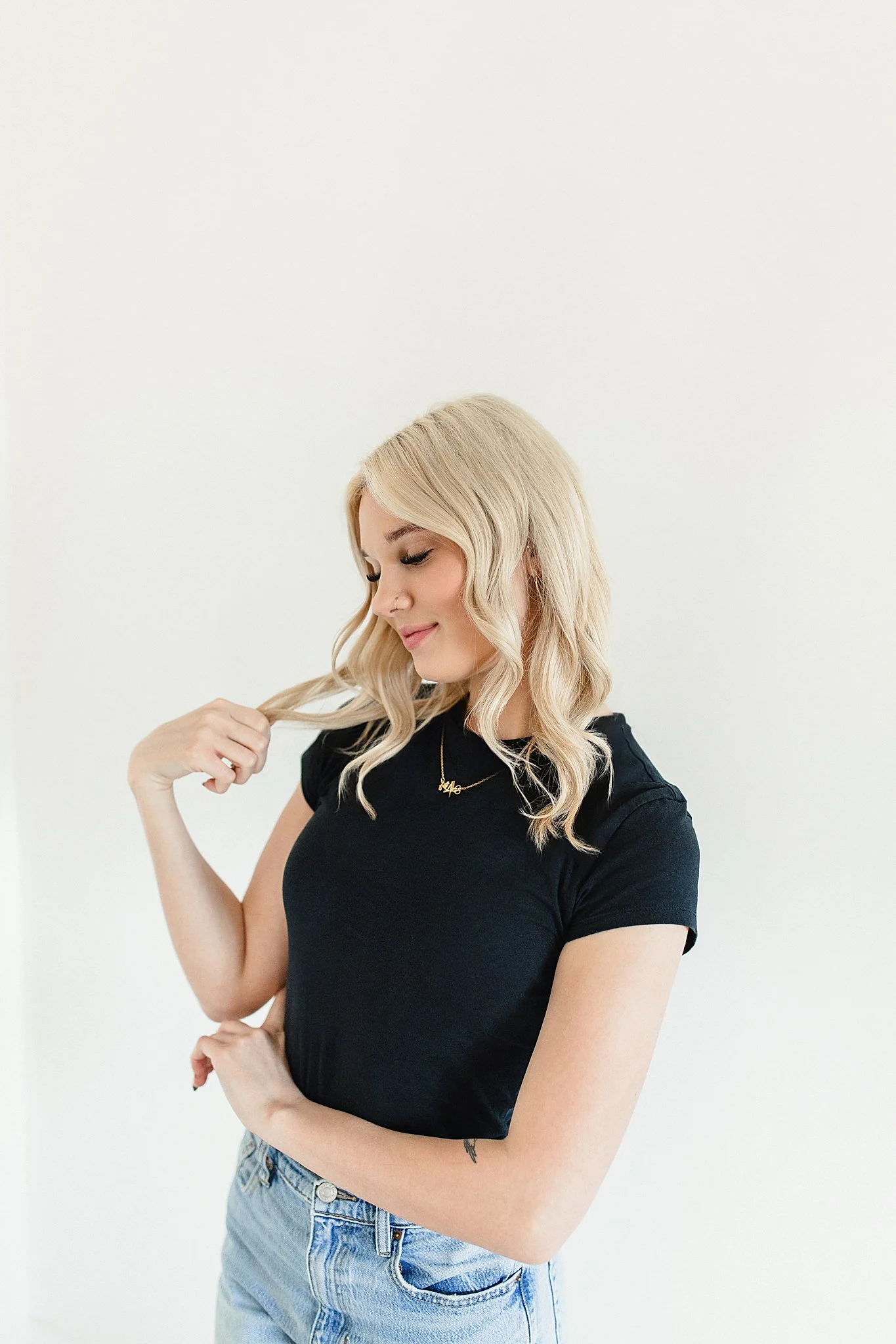 A young woman with blonde hair, wearing a black t-shirt and light blue jeans, is smiling and gently holding a strand of her hair against a white background for small business branding near Naperville, IL.