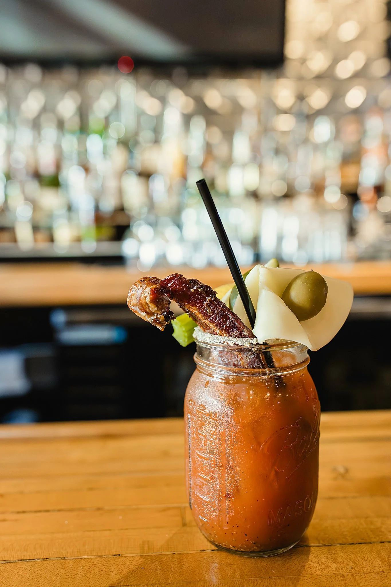 A cocktail in a mason jar glass with a slice of bacon, a green olive, and a cheese cube on top, inside a bar or restaurant with a blurred background for cocktail menu items with commercial photographer Ally and B Photography.