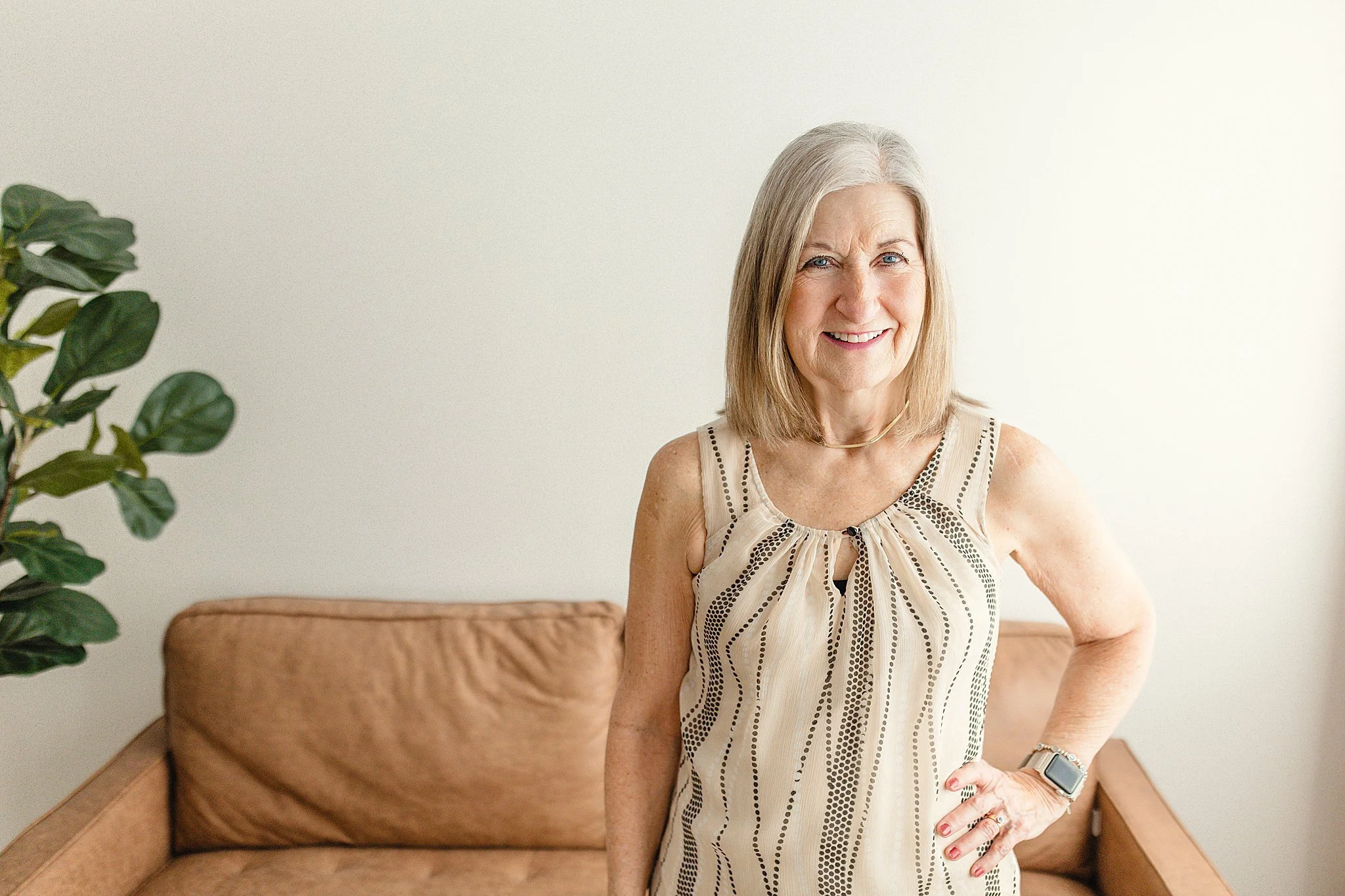 A smiling older woman with shoulder-length blonde hair standing in front of a beige couch, wearing a sleeveless beige dress with black dotted patterns, a necklace, and a smartwatch on her left wrist during headshots in Naperville, IL.