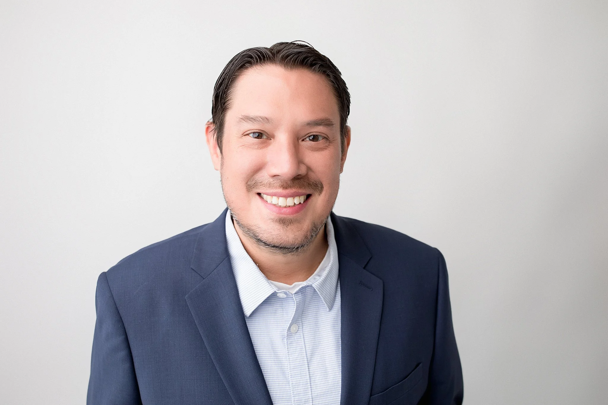 Portrait of a smiling man in a navy blue suit and light blue shirt, against a plain light gray background during a headshot photography session with Ally and B Photography in Naperville, Illinois.