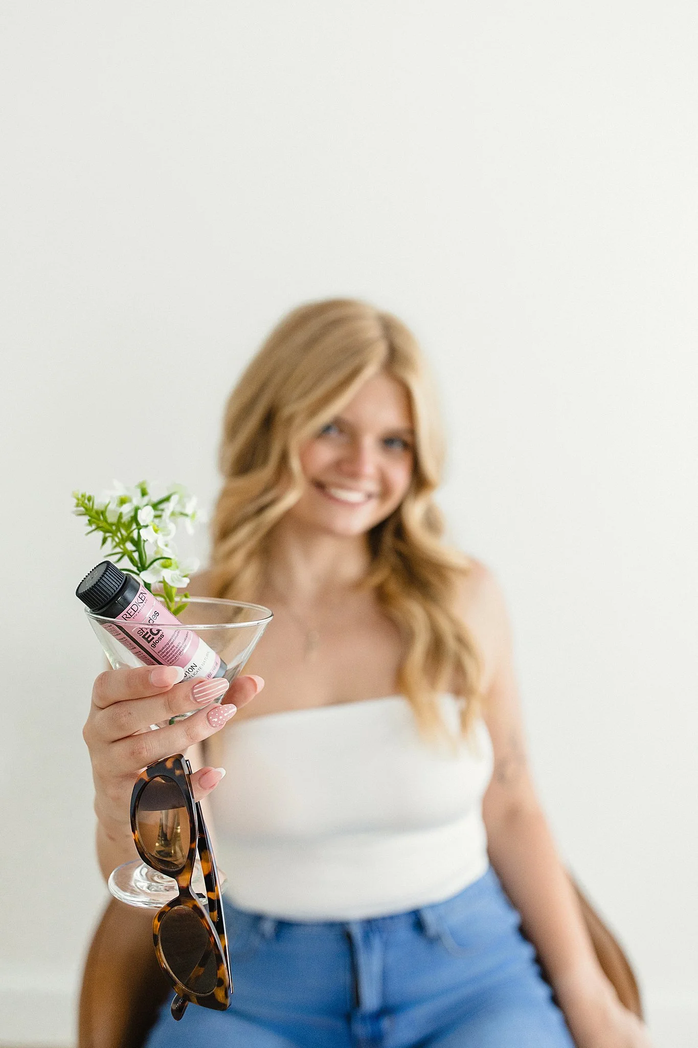 A woman with blonde wavy hair smiling, holding a martini glass with a pink lip gloss tube and white flowers, wearing a white strapless top and blue jeans, sitting in a brown chair, with sunglasses hanging on the glass with a brand photographer.