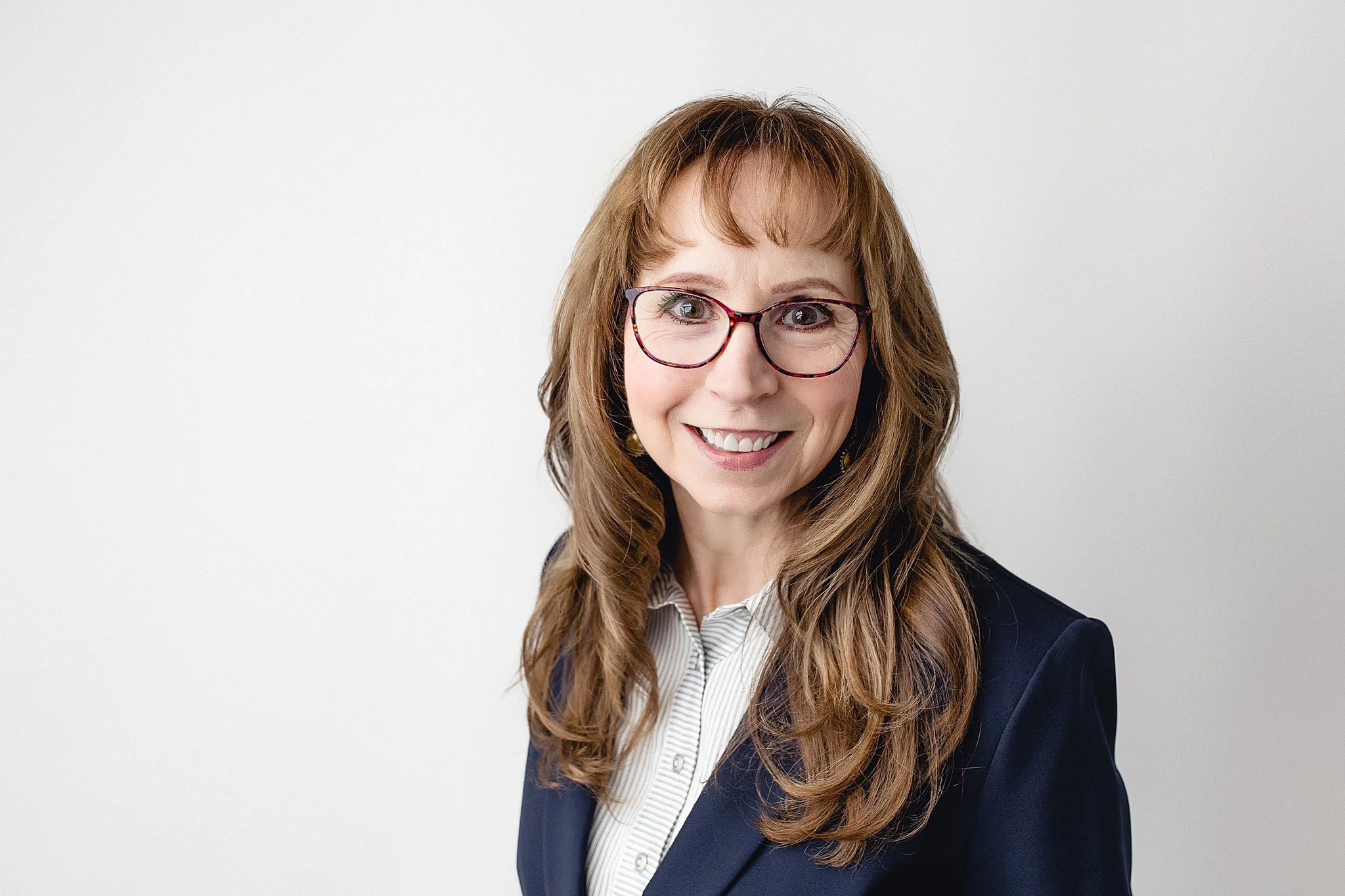 Portrait of a smiling woman with wavy brown hair, glasses, white blouse, and navy blazer against a plain light background with a Naperville headshot photographer.