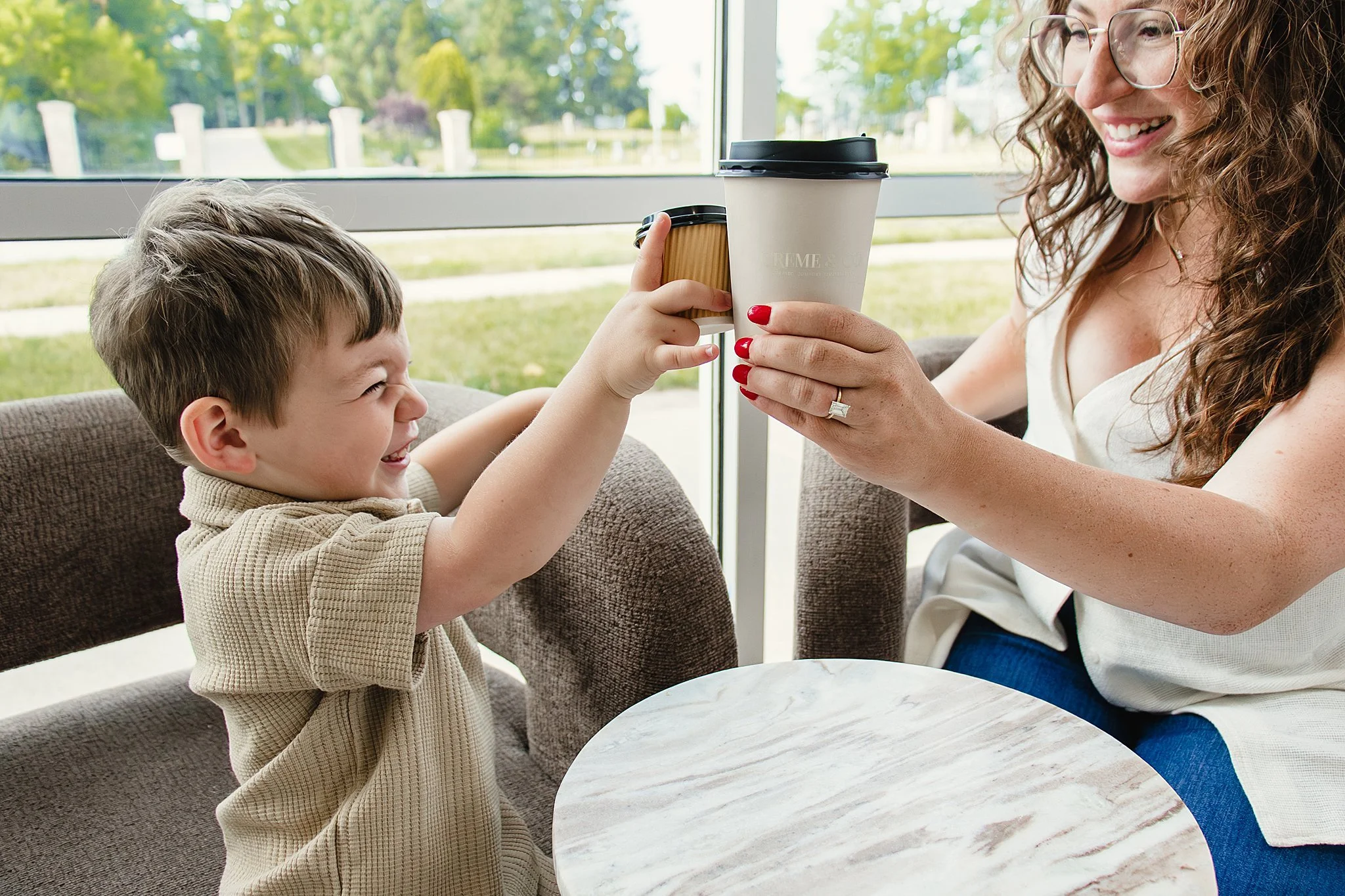 A woman with long, curly hair and glasses handing a paper cup of coffee to a young boy with short hair, both smiling, sitting at a round table near a large window with a view of green trees and a pathway outside for brand photos.