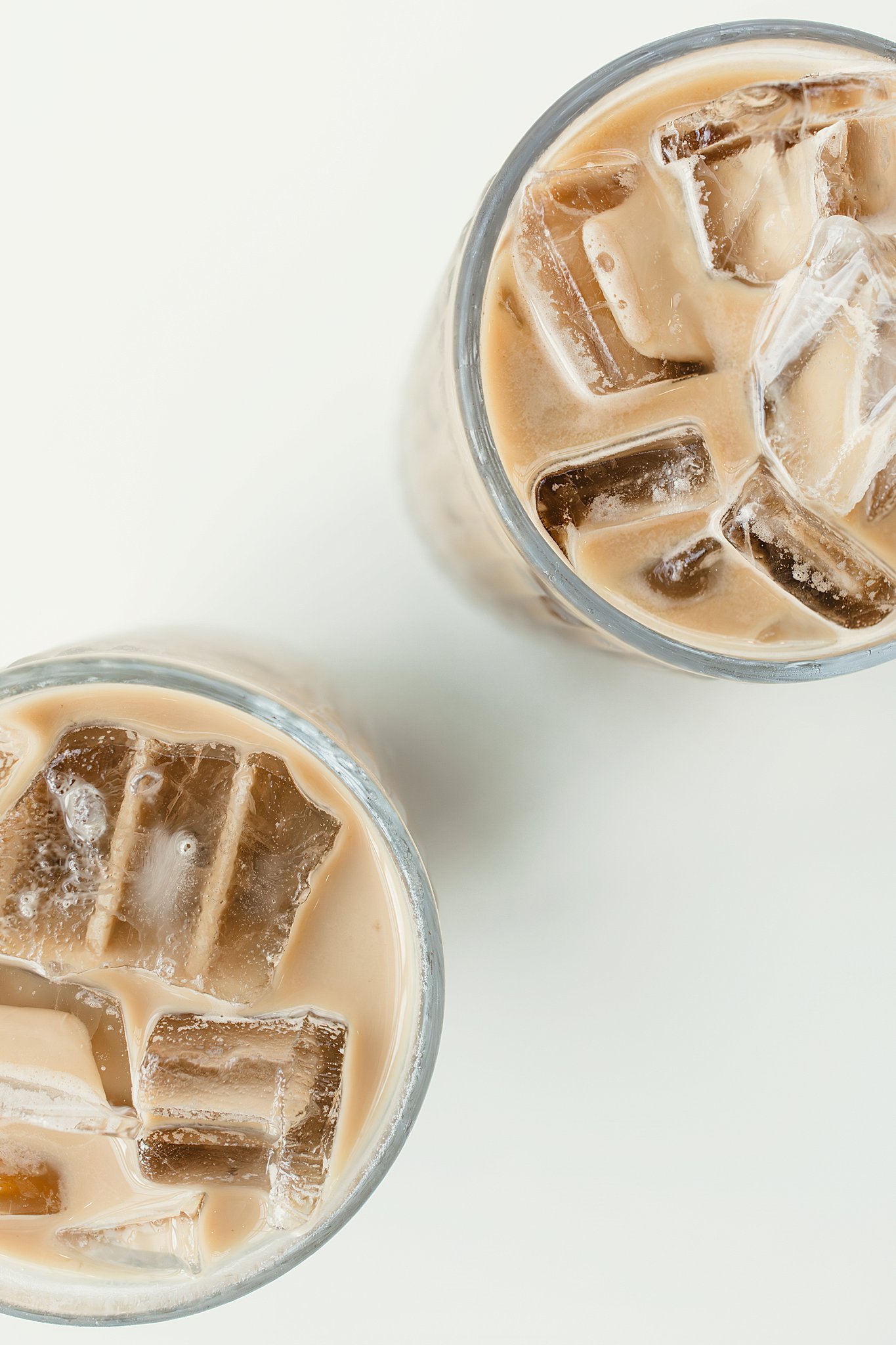 A top view of two glasses filled with iced coffee with milk, served over ice cubes, on a white surface for product photography near me.