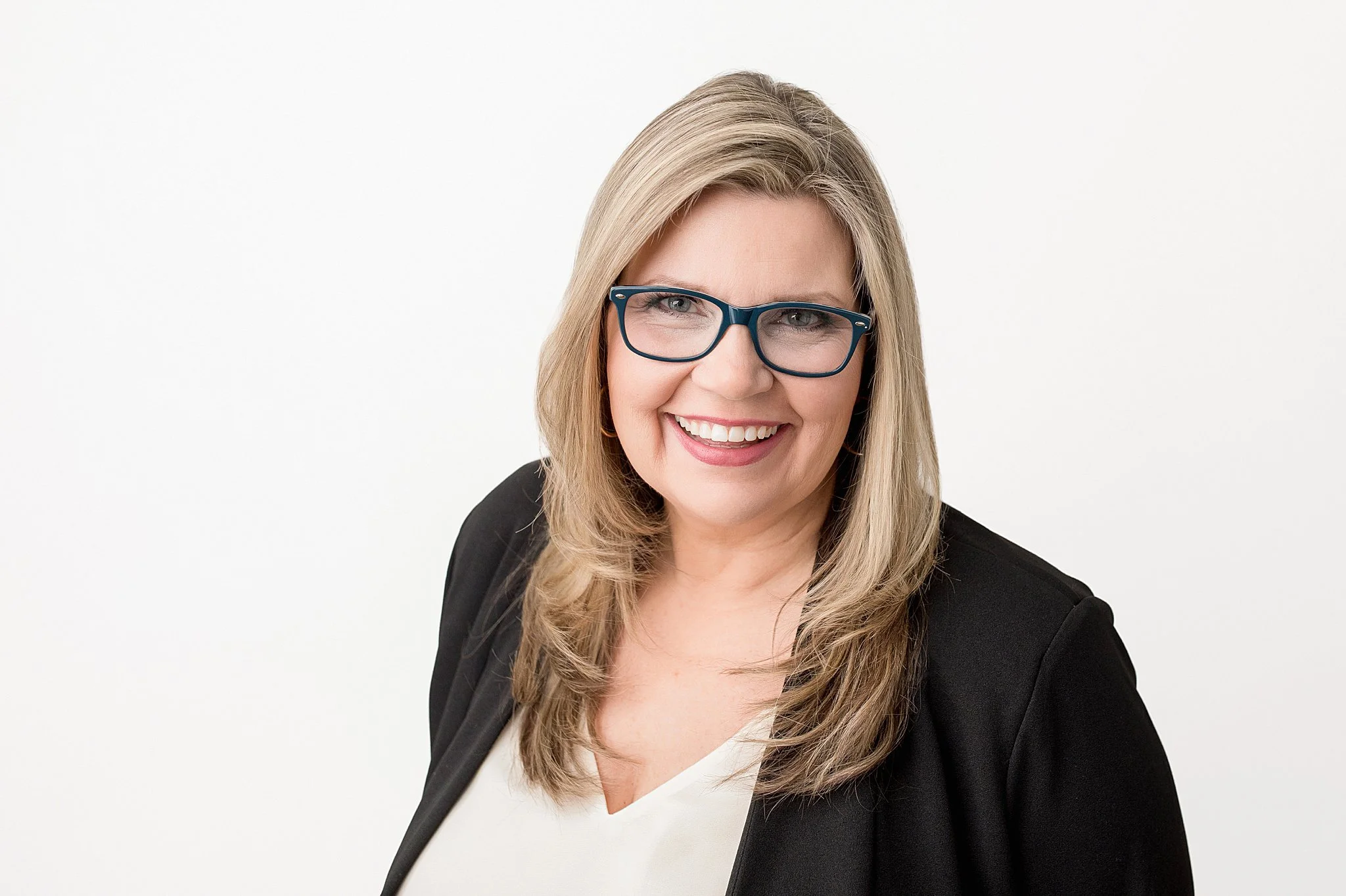 A smiling woman with shoulder-length blonde hair and blue glasses, wearing a black blazer and a white top, against a plain white background with Marie of Ally and B Photography, Naperville headshot photographer.