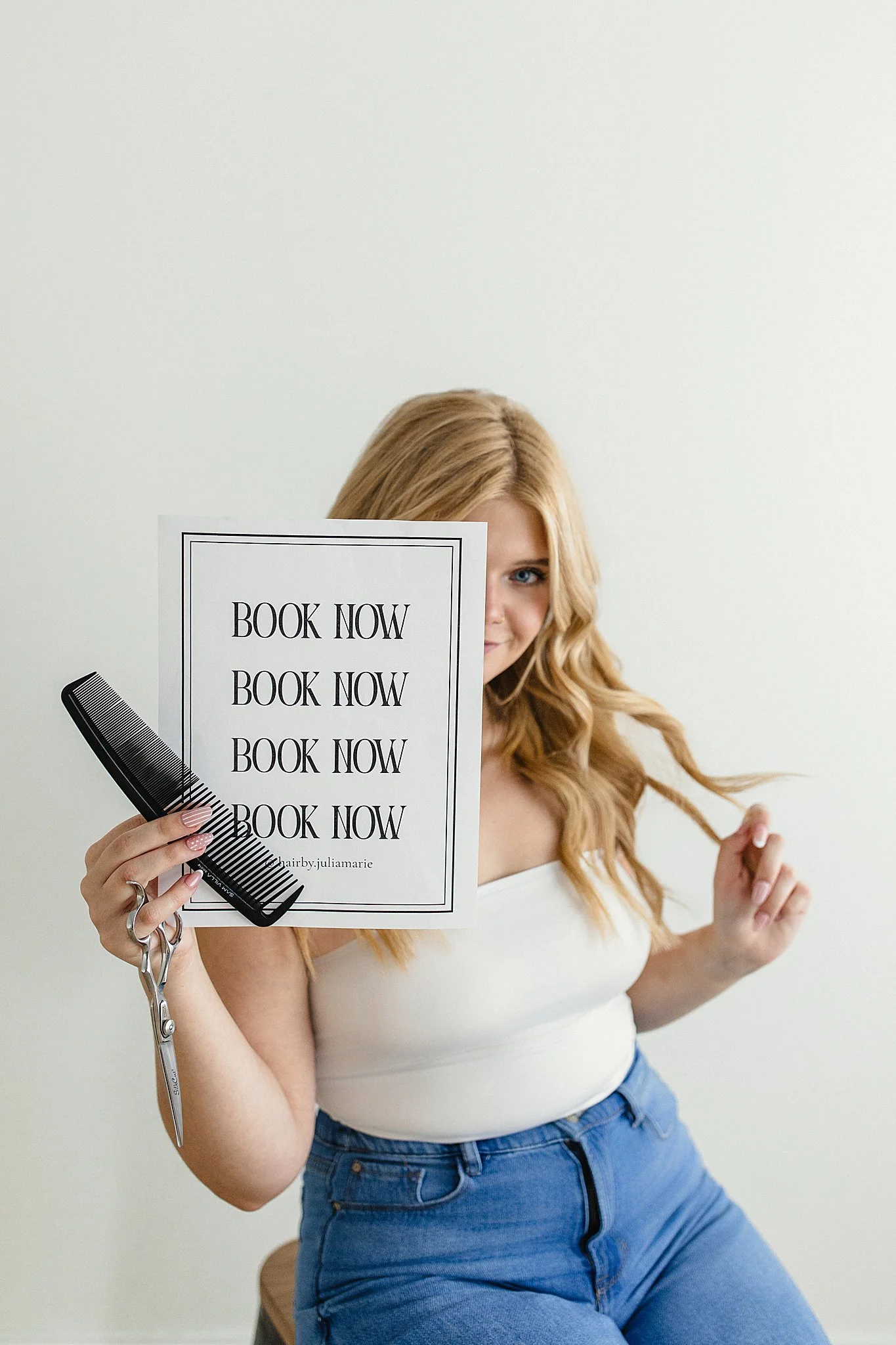 A young woman with long blonde hair, wearing a white top and blue jeans, sitting on a wooden stool, holding a large white paper with black text that says 'BOOK NOW' repeatedly, while also holding a black comb and scissors, with her face partly covere