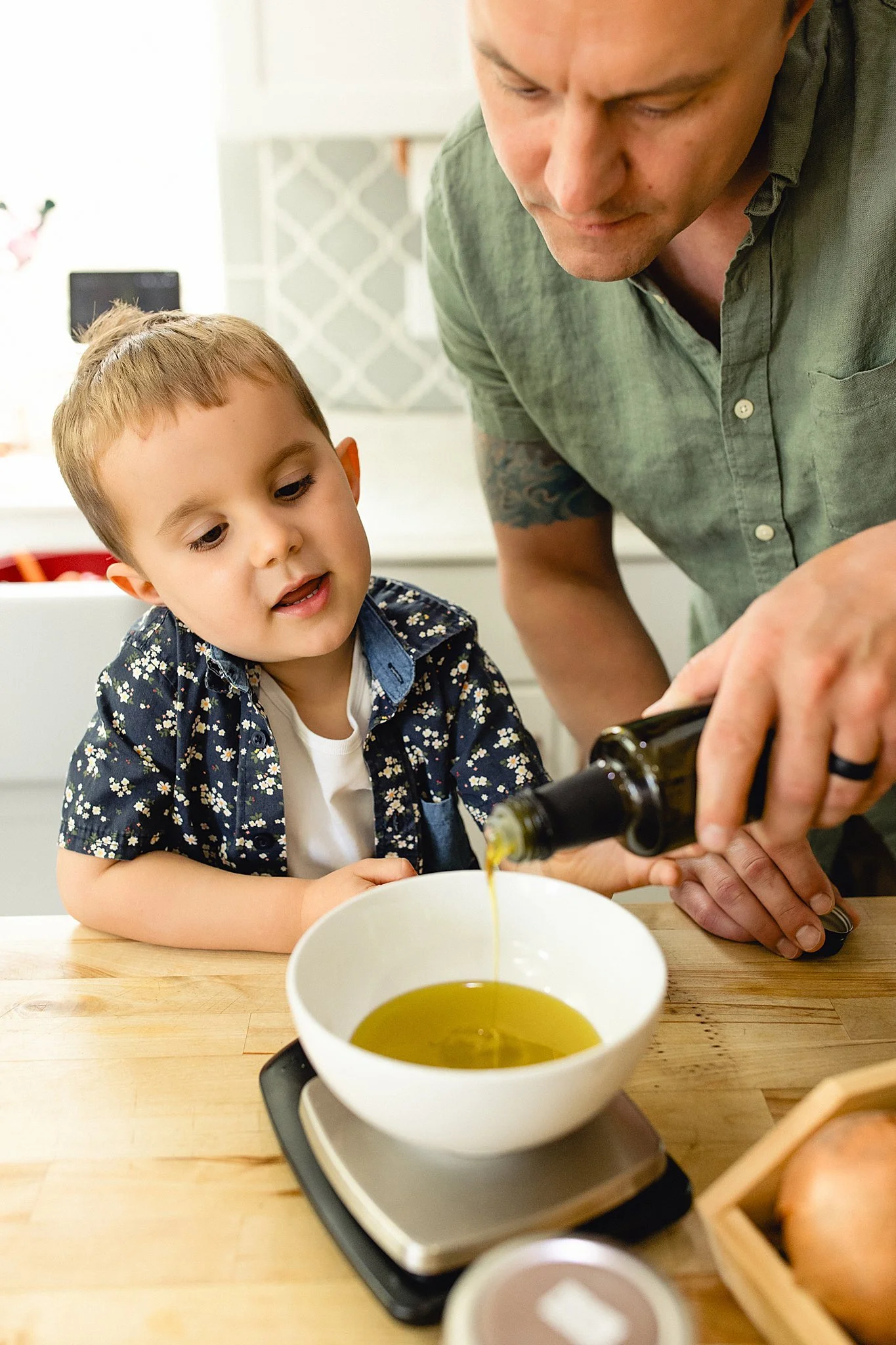 A man pouring olive oil into a white bowl on a kitchen counter while a young boy watches for small business branding photos with Marie Lopez of Ally and B Photography in Naperville, IL.