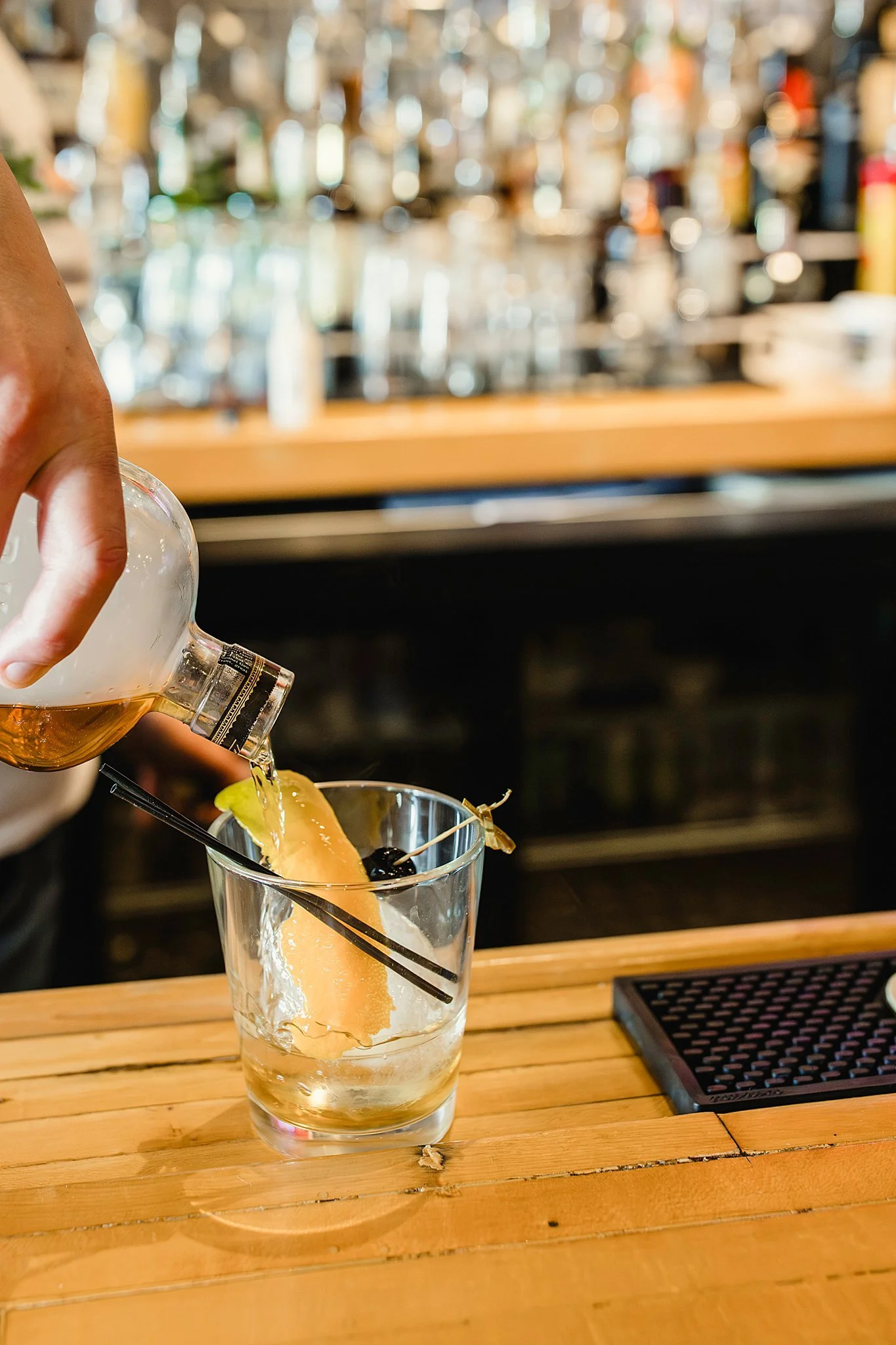 Person pouring a drink into a glass with ice and garnish at a bar counter during a bar commercial photography session for cocktail menu items.