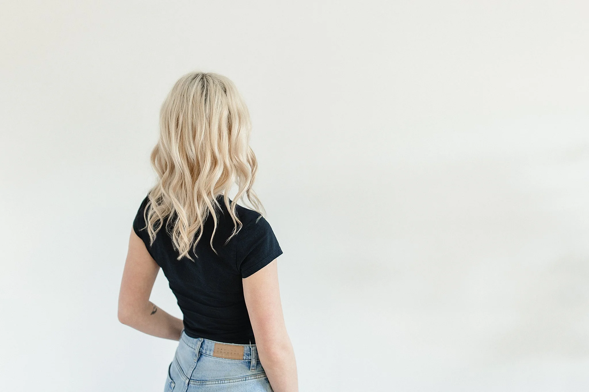 Back view of a woman with blonde wavy hair, wearing a black t-shirt and light blue jeans, standing against a plain white wall for small business branding near Naperville.