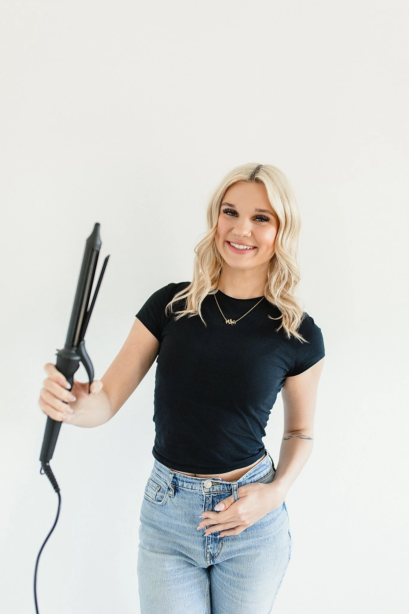 Young woman with blonde hair smiling, holding a curling iron, wearing a black t-shirt and jeans, standing against a plain white background during hairstylist branding photos in Naperville, IL.