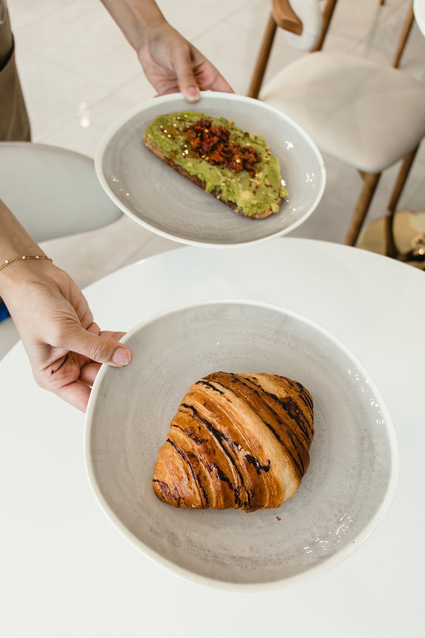 A croissant with chocolate drizzle in a white bowl, and a slice of bread with avocado and toppings on a plate, being held by a person over a white table with branding photographers near me.