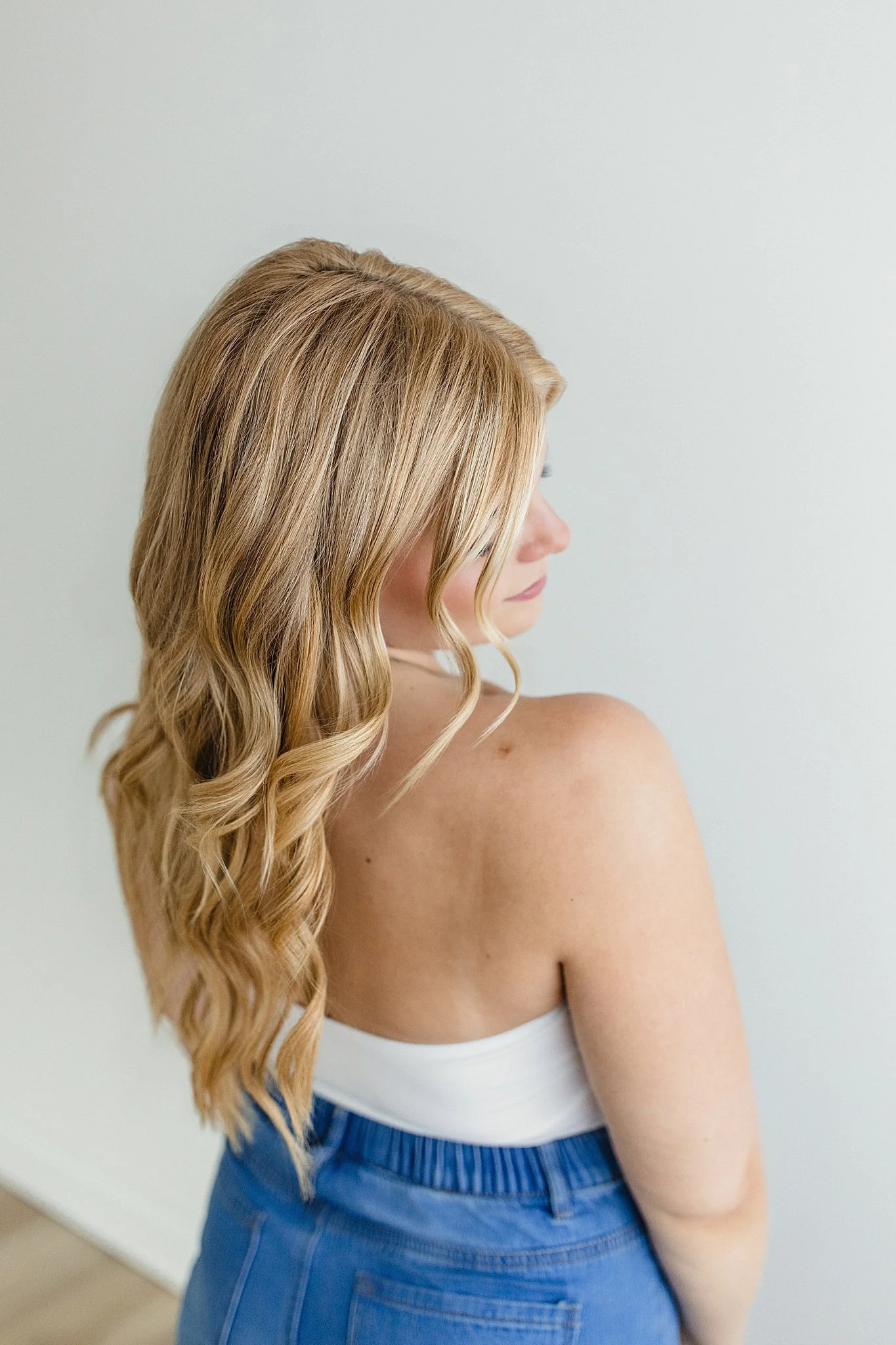 A woman with long, wavy blonde hair seen from the side, wearing a white tank top and blue high-waisted jeans, standing against a plain light-colored wall with branding photographers near me.