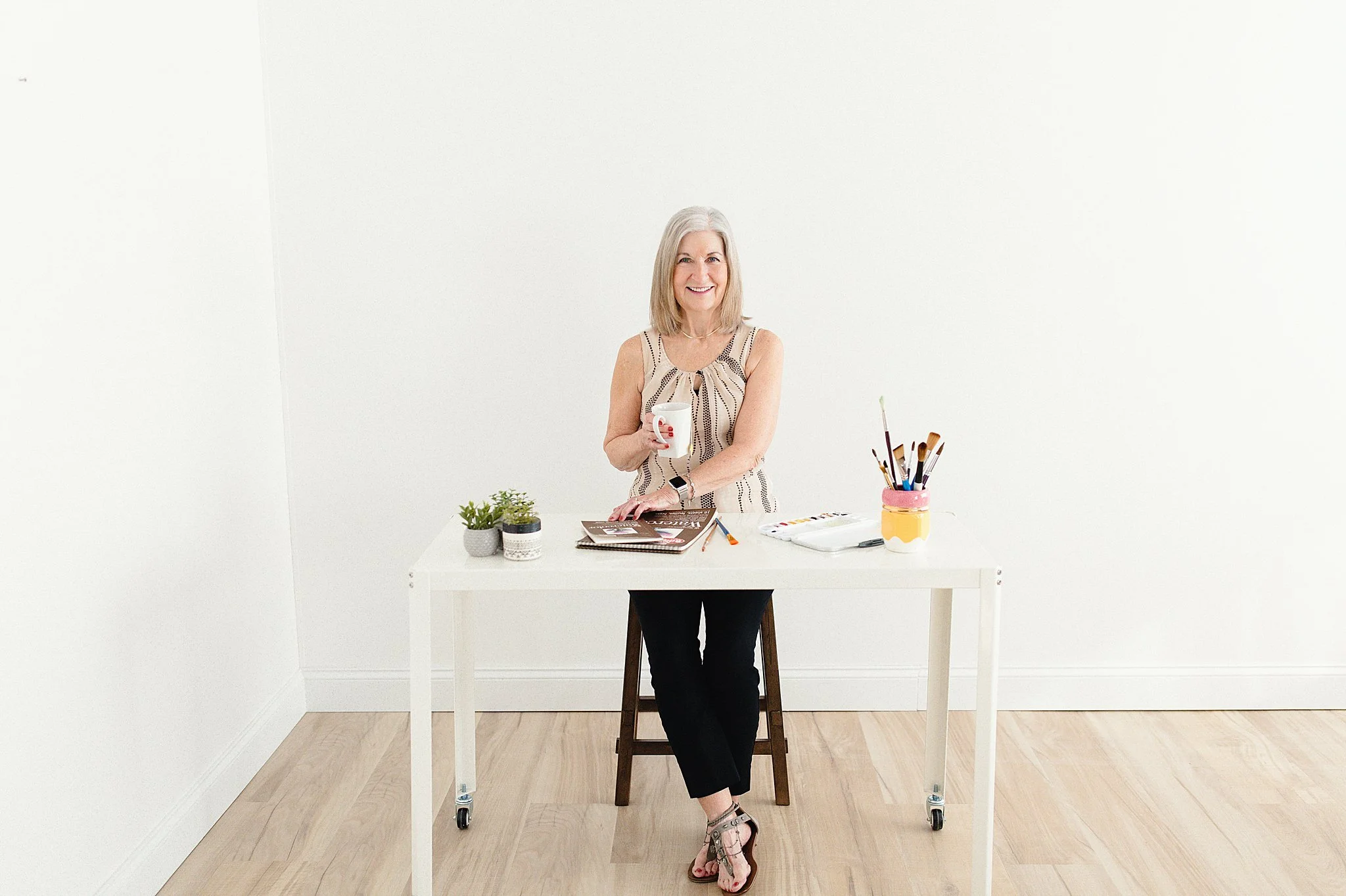 A smiling woman with gray hair sitting at a white table holding a white mug in her right hand, with art supplies and small potted plants on the table, in a minimalistic room with white walls and wooden floor during branding photography near me.