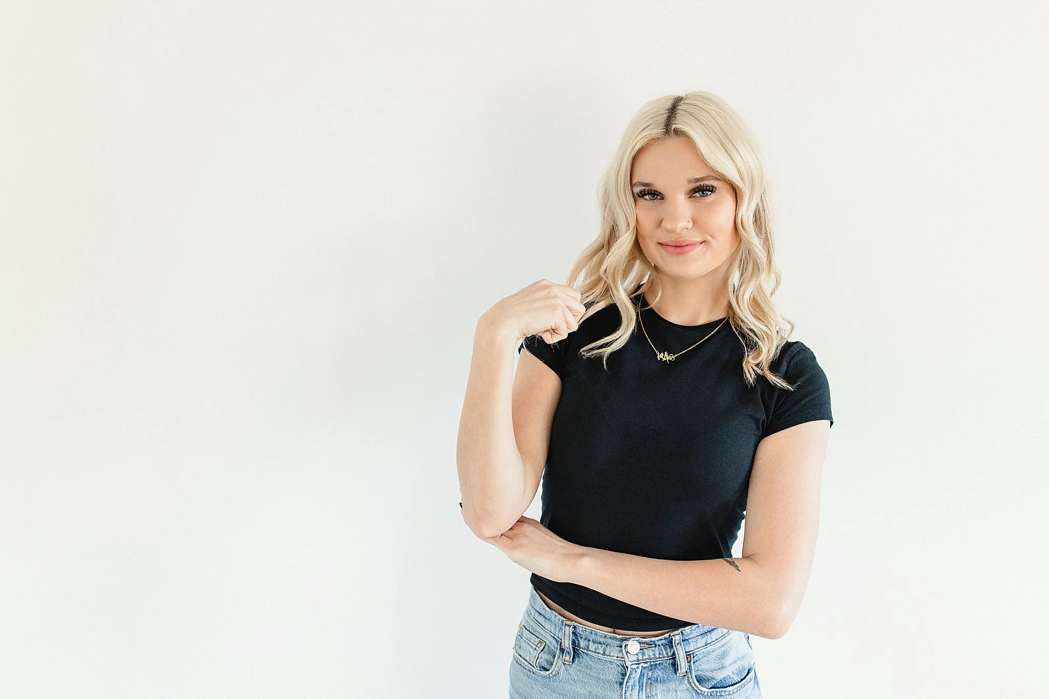 A young woman with blonde wavy hair, wearing a black T-shirt and light blue jeans, smiling softly against a plain white background for a brand photoshoot in Naperville, IL.