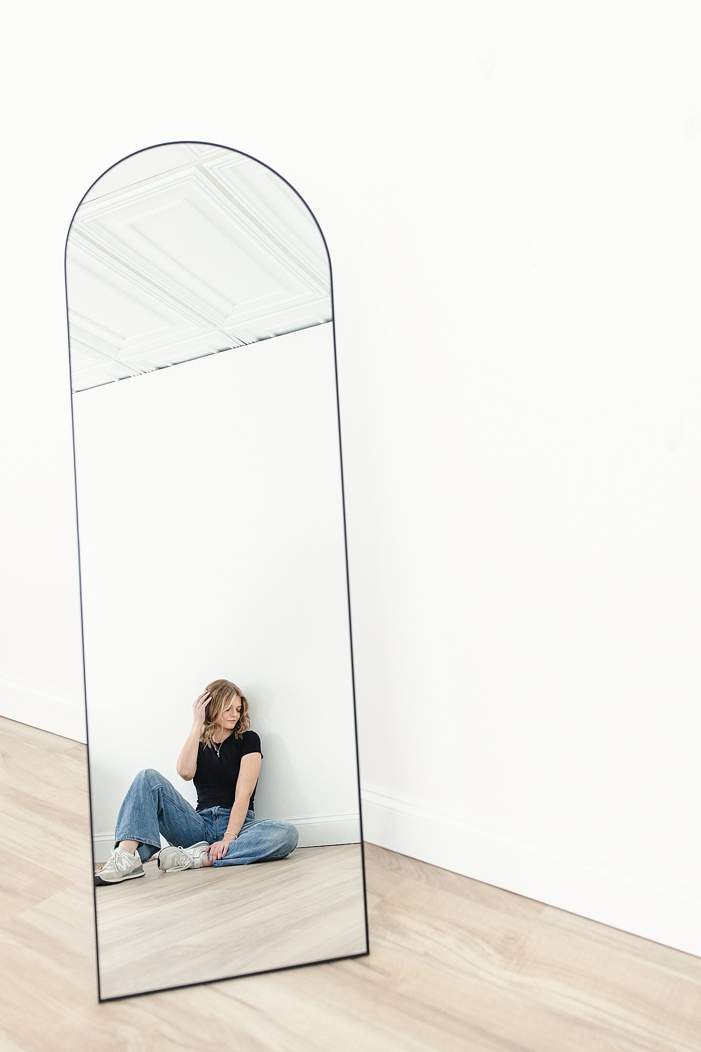 A woman sitting on the floor looking at her reflection in a tall, arched mirror against a white wall, with a wooden floor during a hairstylist brand photography session with Ally and B Photography in Naperville, IL.