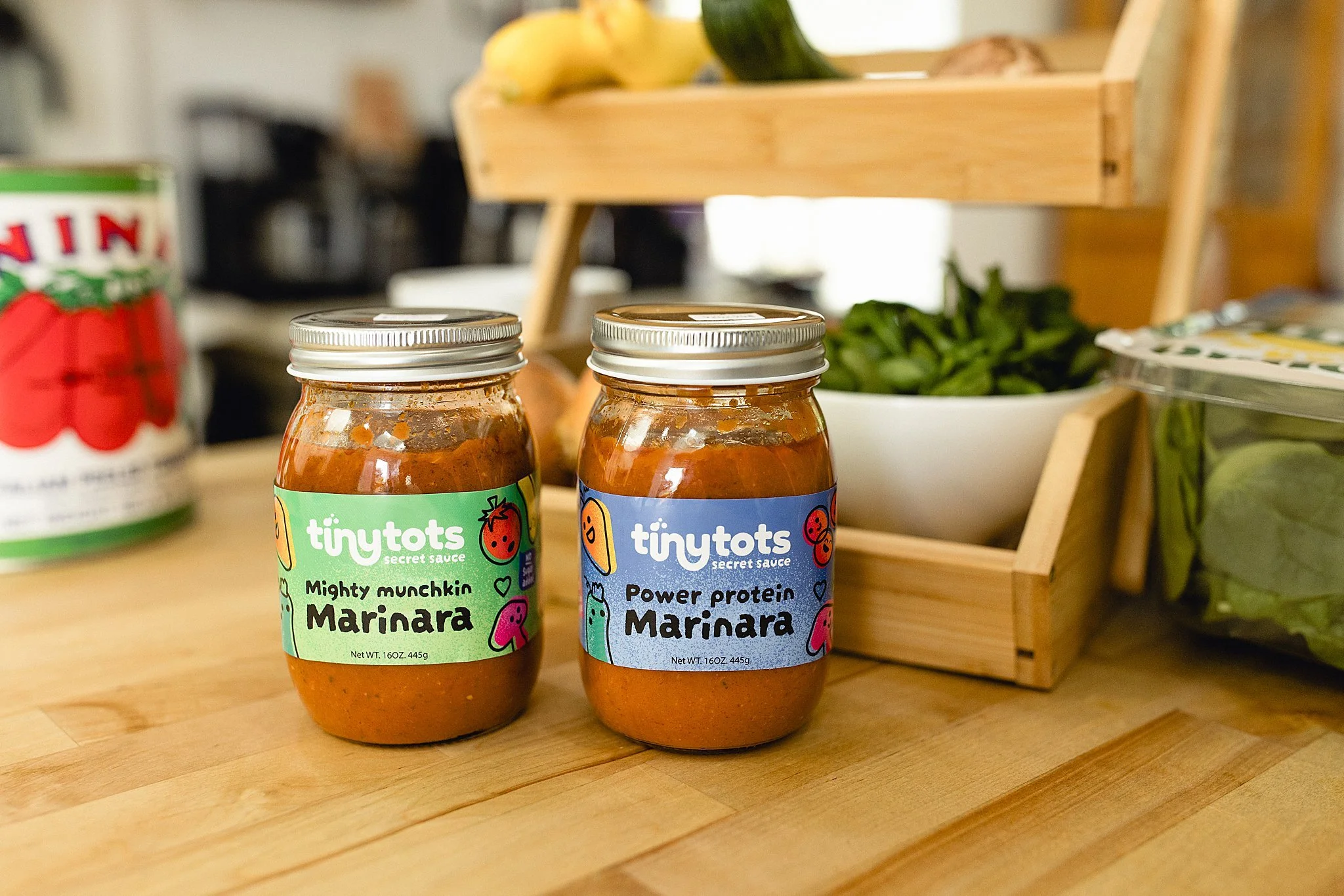Two jars of Tiny Tot's secret sauce on a wooden surface, labeled Mighty Munchkin Marinara and Power Protein Marinara, with fresh spinach in a bowl and other grocery items around for product photography in Naperville, IL.